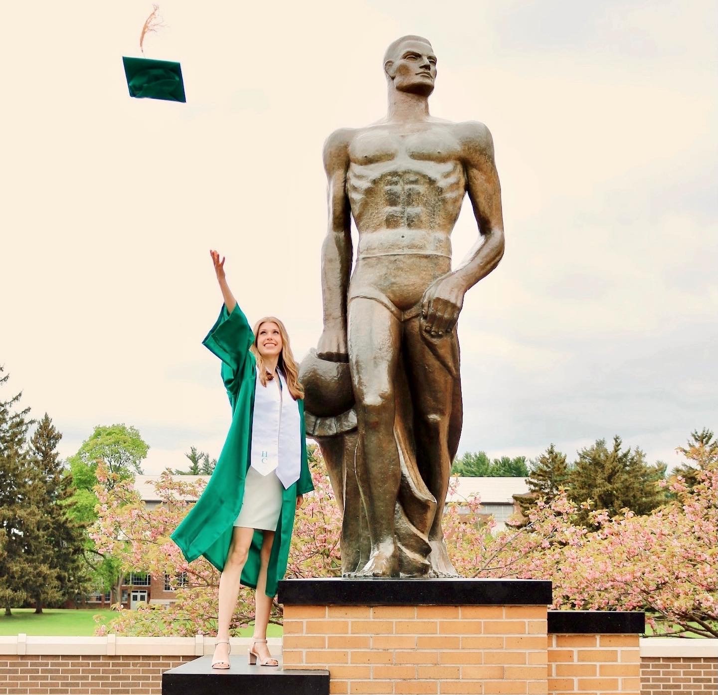 Allie Virginski in graduation gown, throwing cap into the air, while standing atop the Spartan statue