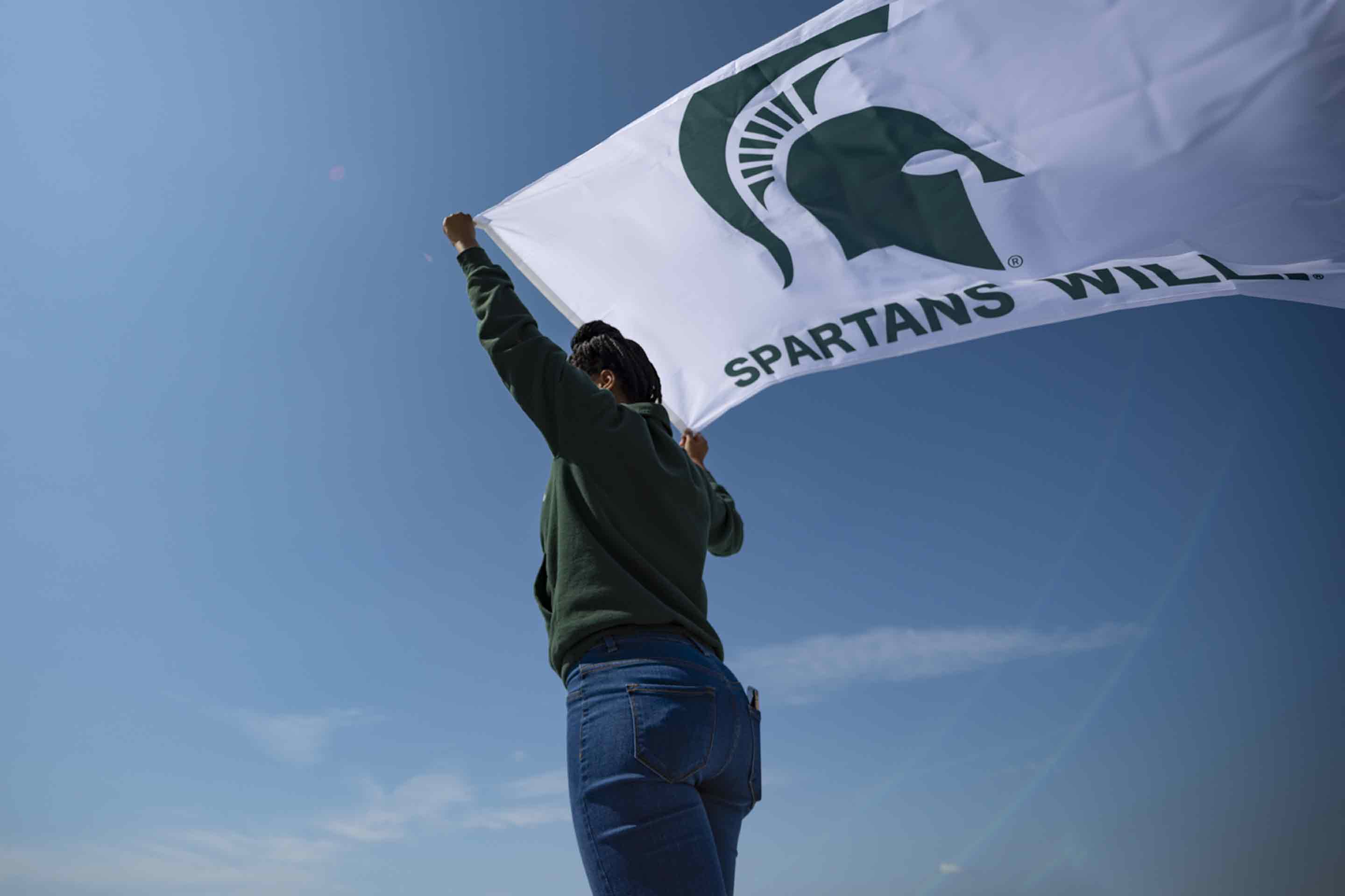 Spartan holding MSU flag with a blue sky as the back drop. 