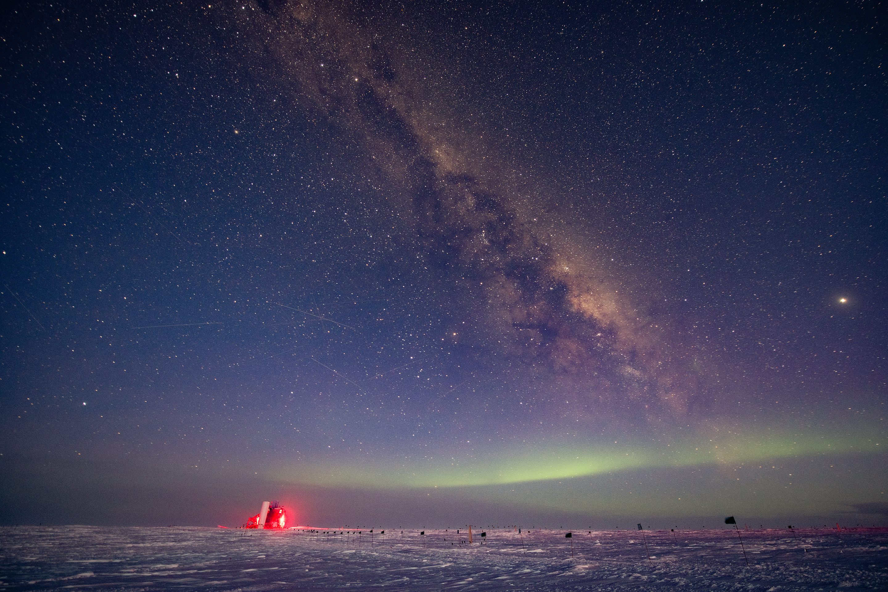 The IceCube Laboratory, lighted red against the night sky, is small in this landscape photograph of the South Pole white tundra, which also captures yellow stars and light green auroras.