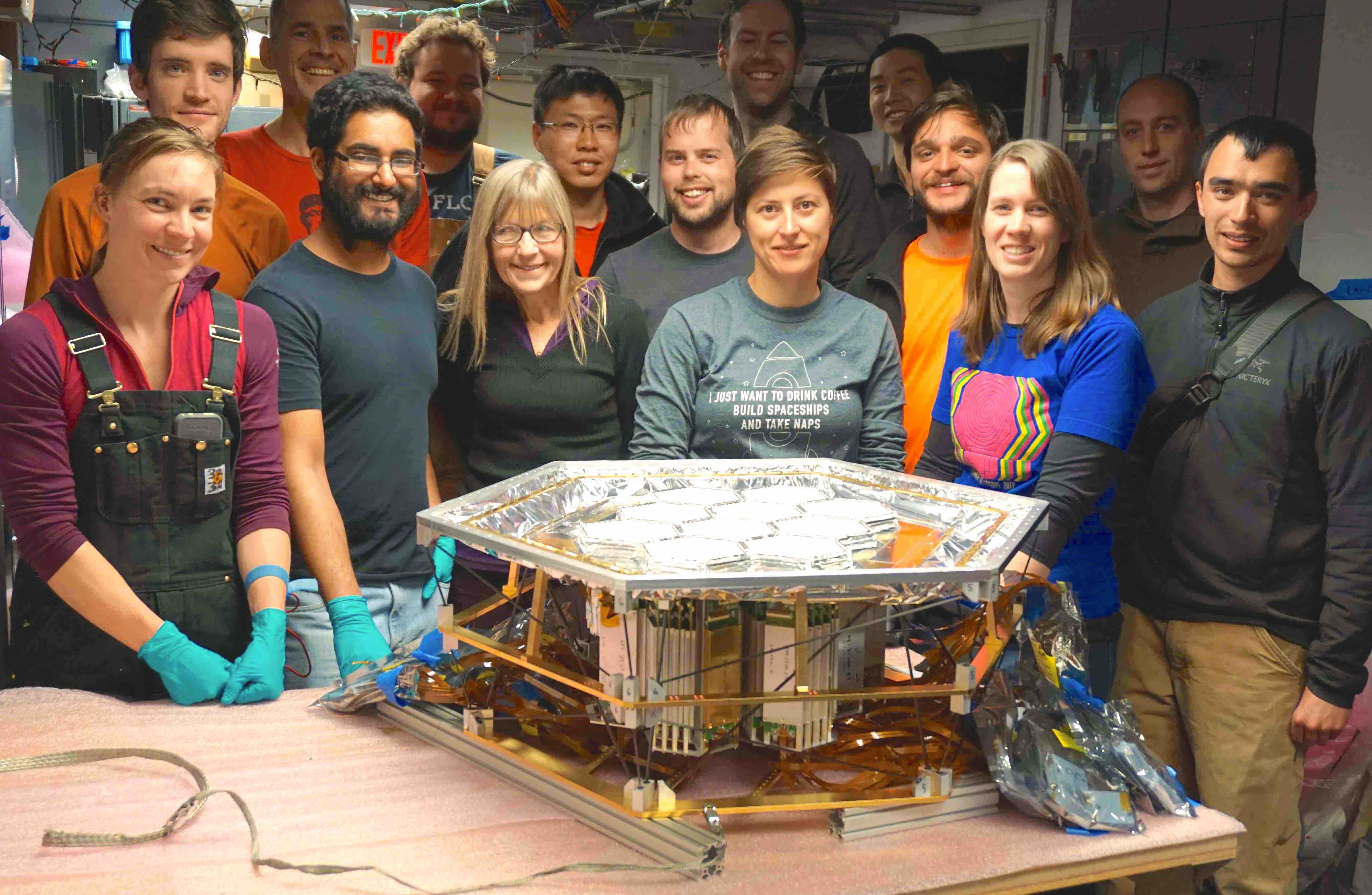 Several scientists stand around a large, silvery hexagonal instrument sitting on a table.