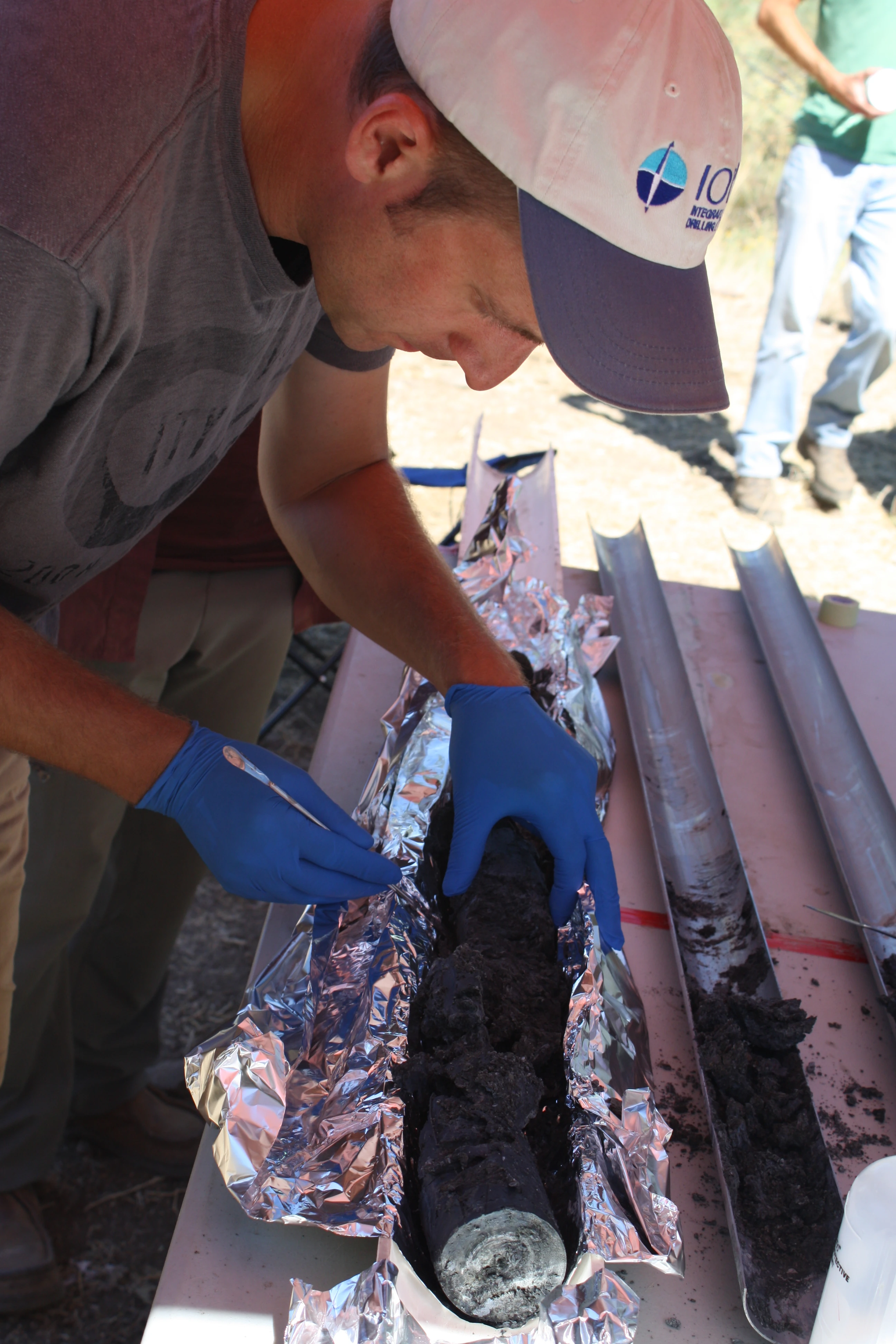 MSU Associate Professor Matthew Schrenk, wearing blue gloves, examines a dark cylindrical sample outdoors at a research site.