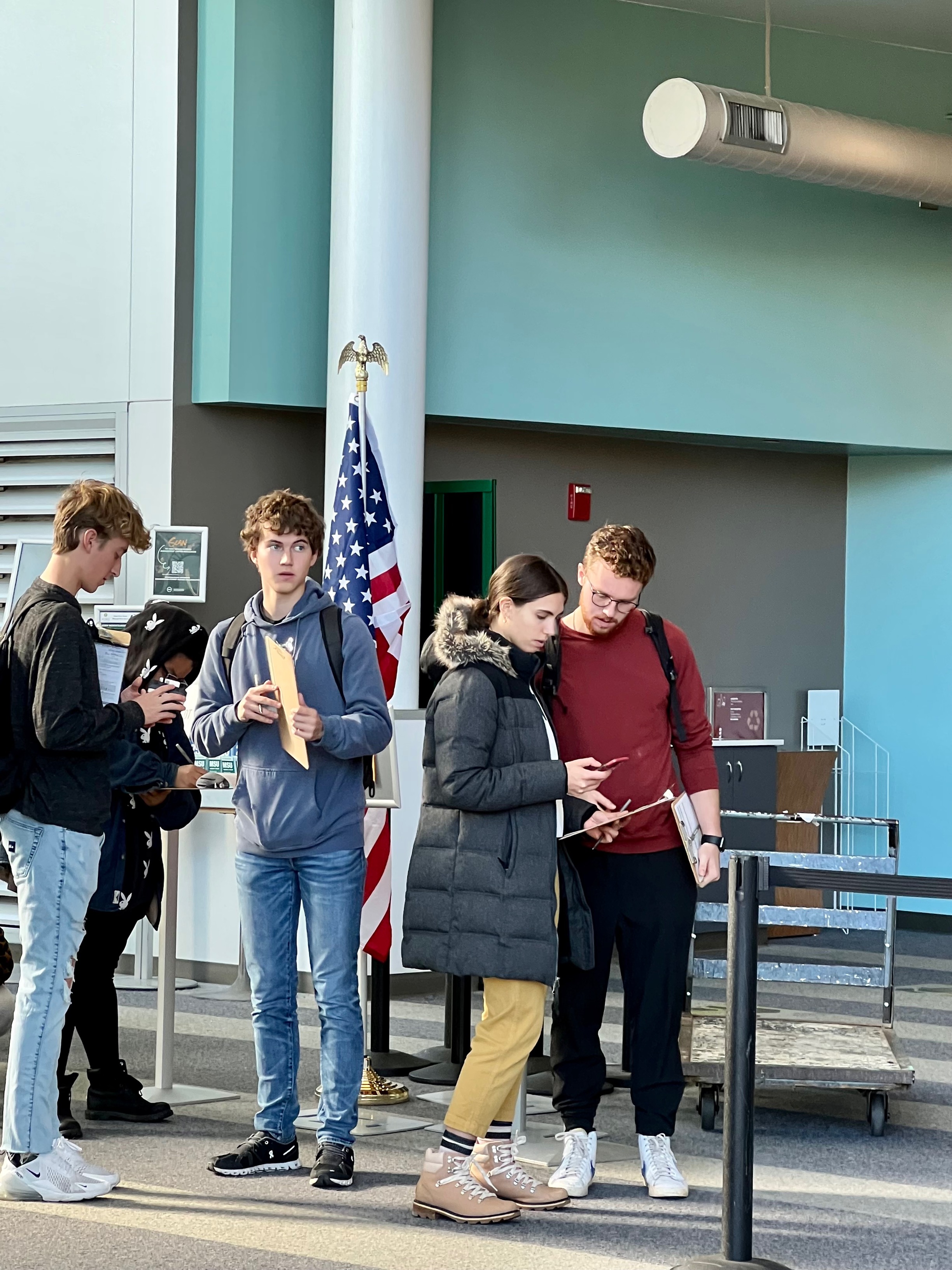 Four students waiting in line to vote in the 2022 midterm elections with an American flag in the background.