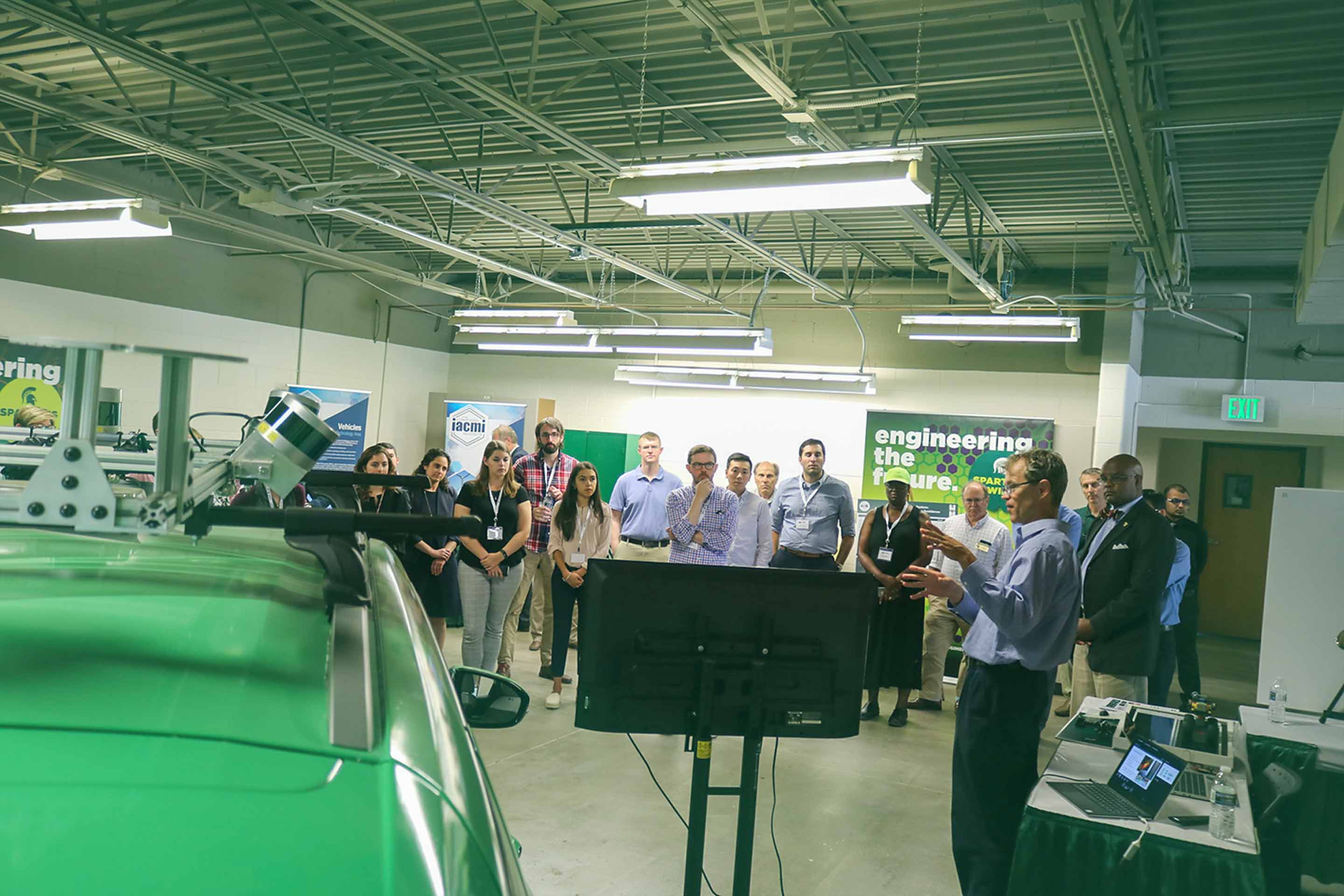 MSU Associate Professor Daniel Morris stands in front of a group in a lab space with one of MSU’s autonomous vehicles.