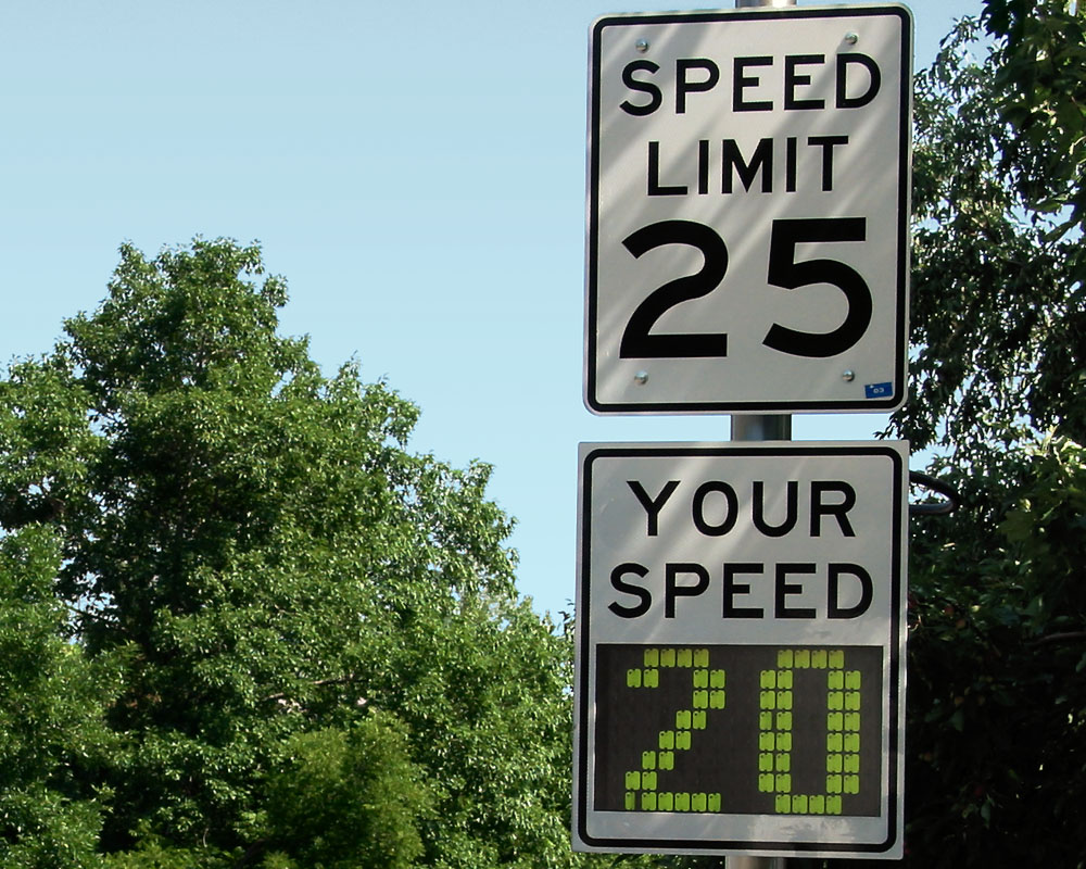 Two white speed limit signs are aligned vertically and seen against a blue sky in front of green trees. The top sign says “SPEED LIMIT 25” in black text. The lower sign reads “YOUR SPEED” in black and a green LED display shows “20.”