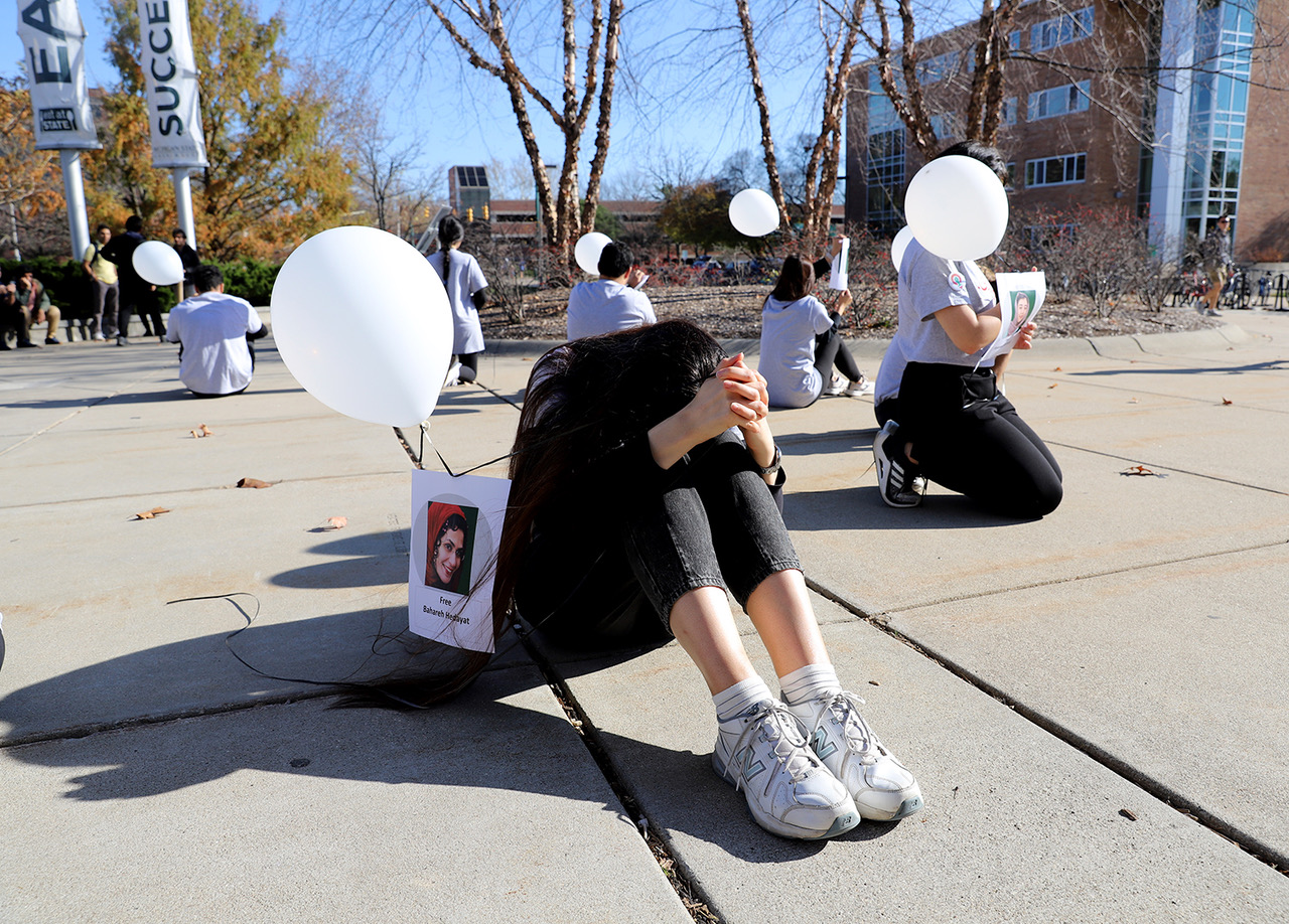 Performance by Parisa Ghaderi for Iran prisoners and detainees featuring performers sitting on the ground outdoors holding a white balloon tied to a printed page featuring a detainee.