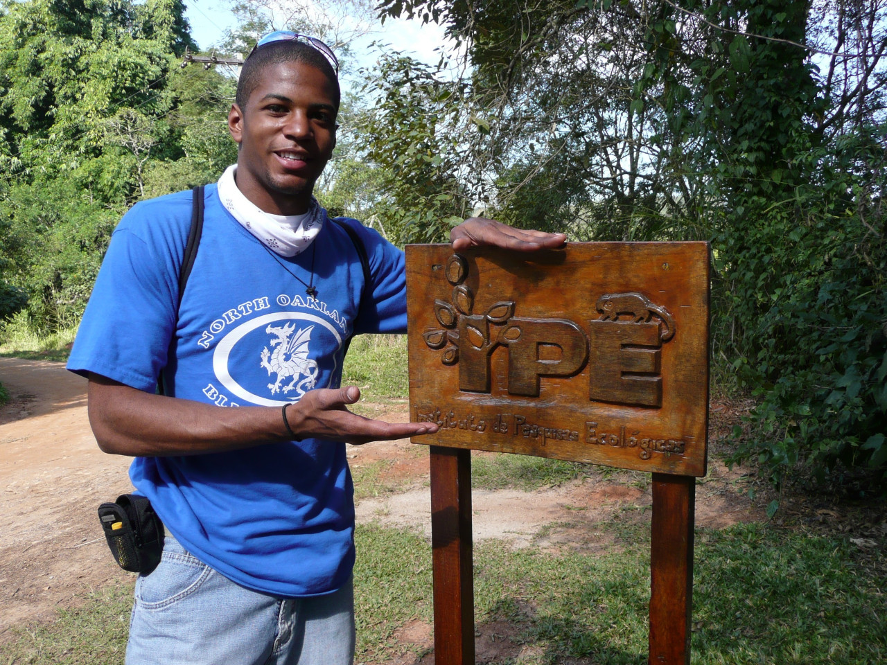 A Black man in a blue t-shirt stands next to a brown wooden sign.