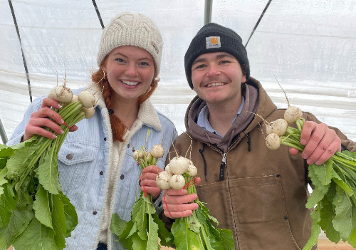 A female student on the left and male student on the right smile at the camera. They are standing in a greenhouse holding turnips.