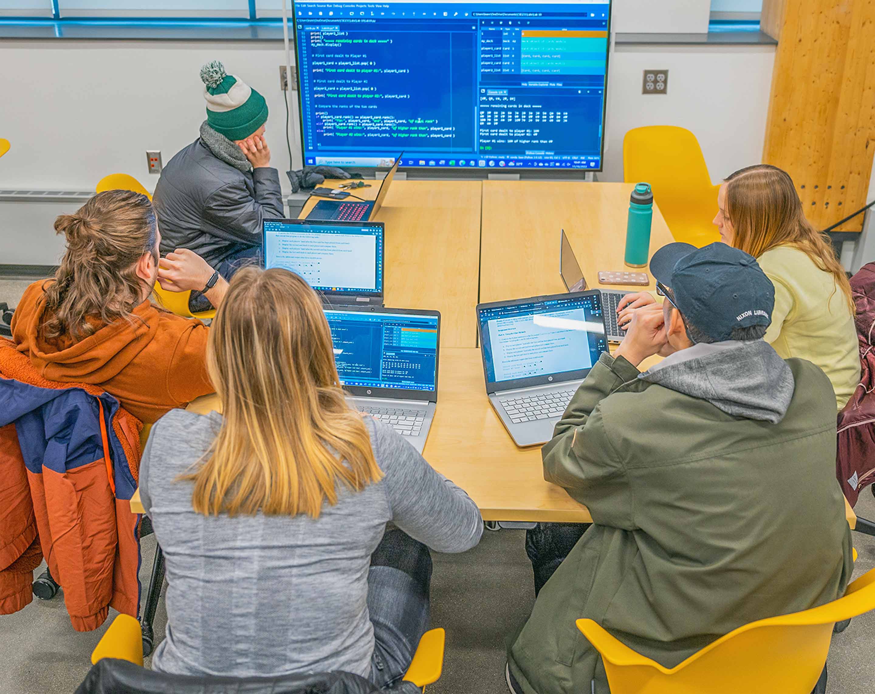 Students sitting at a table with laptops review  code on a large monitor during an introduction to programming class.