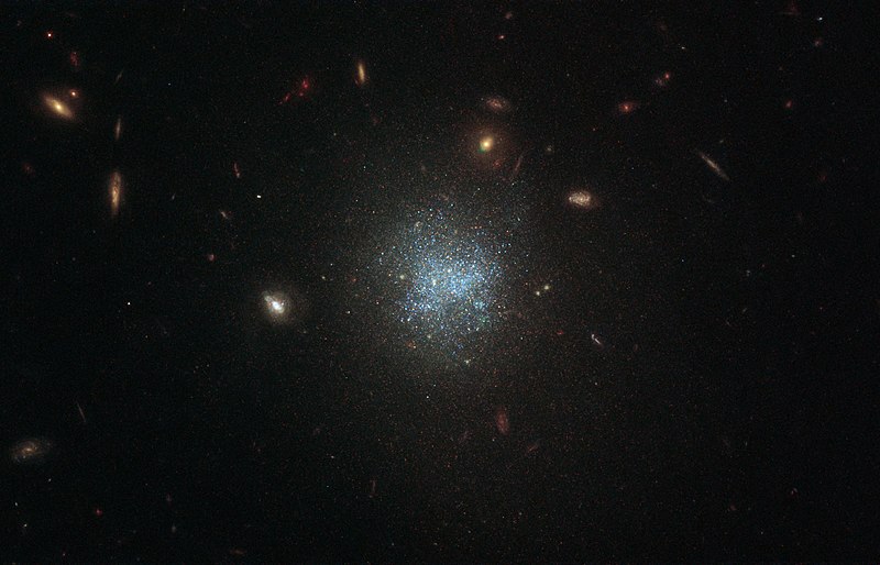 A dark telescope image is dotted with small yellow, disc-shaped spiral galaxies.  At its center, however, the galaxy depicted looks like a scattered cloud of blue dots.