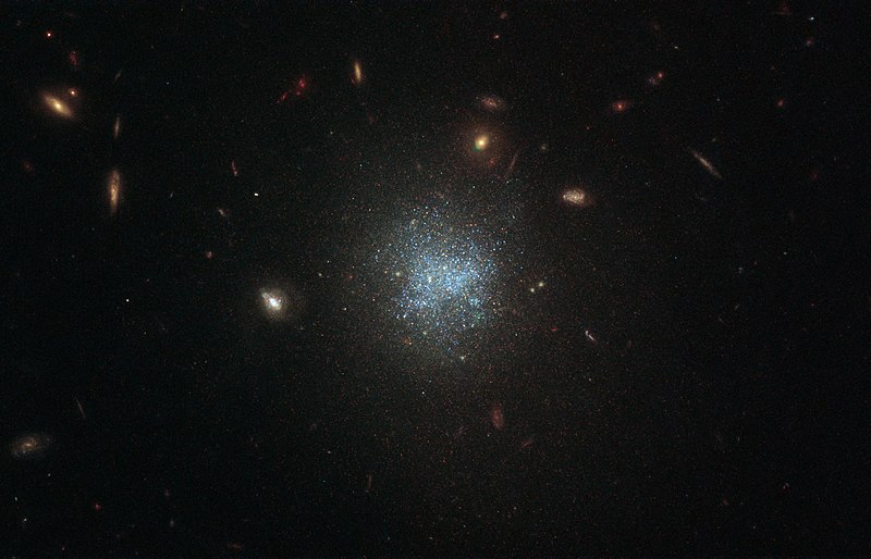 The dark telescope image is dotted with small yellow spirals and disk-shaped galaxies. But the galaxy in its center looks like a scattered cloud of blue dots.