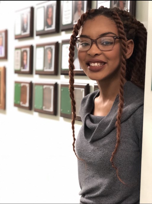Similing Black woman in glasses and a gray shirt standing in front of a white wall with frames. 