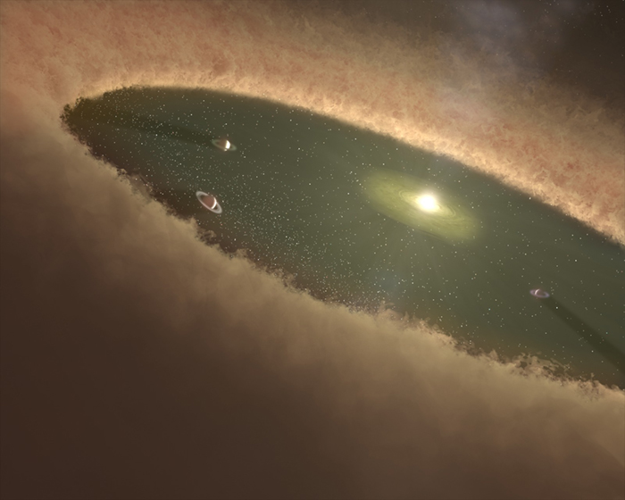 An artist’s rendering shows a hypothetical early solar system with a bright yellow star making a hole in light brown cloud of gas and dust that stretches across the solar system. In the cleared area, three gas giant planets with rings surround the star.