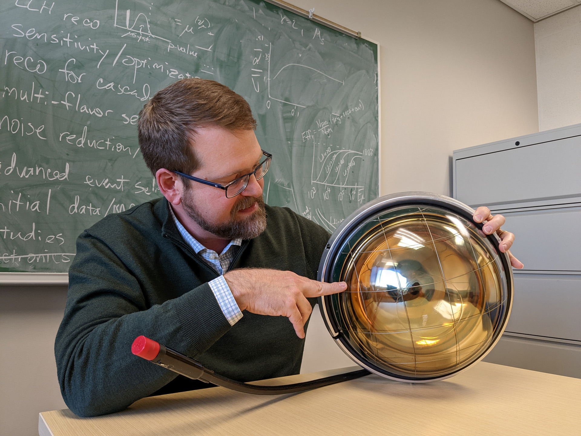 MSU Professor Tyce DeYoung sits in front of a green chalkboard covered with notes, graphs and equations. He’s holding a metal and glass gold orb, which is a digital optical module that IceCube uses to detect neutrinos.