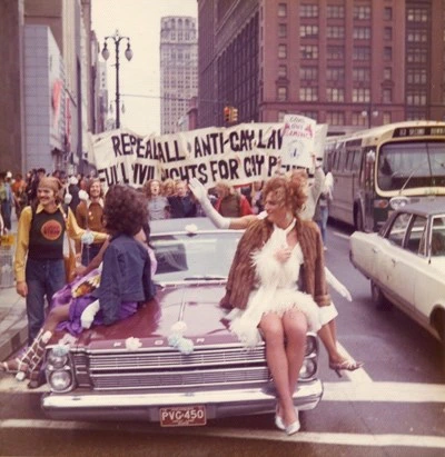 Miss Capital City riding on the hood of a car wearing a white dress at the Christopher Street Detroit '72 Pride march with other parade participants waving gleefully.
