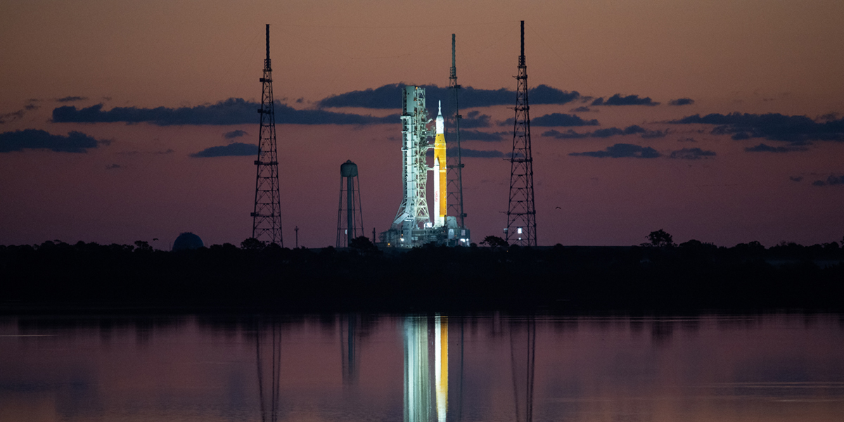 The Artemis I launch stage is illuminated at dawn at Kennedy Space Center in Florida. NASA’s white Orion spacecraft is fixed to the top of the tall, cylindrical orange and white Space Launch System rocket.