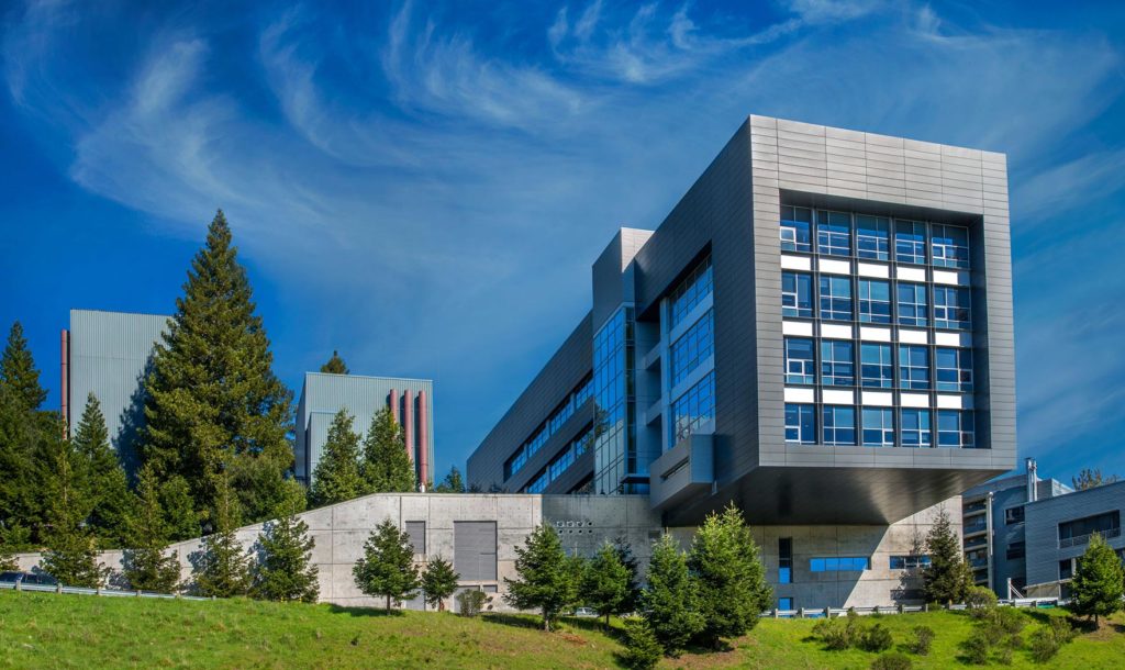 A photograph of the Molecular Foundry — a rectangular charcoal-colored building with mosaic-like blue windows — stands among green pines and against a blue sky with wispy white clouds.