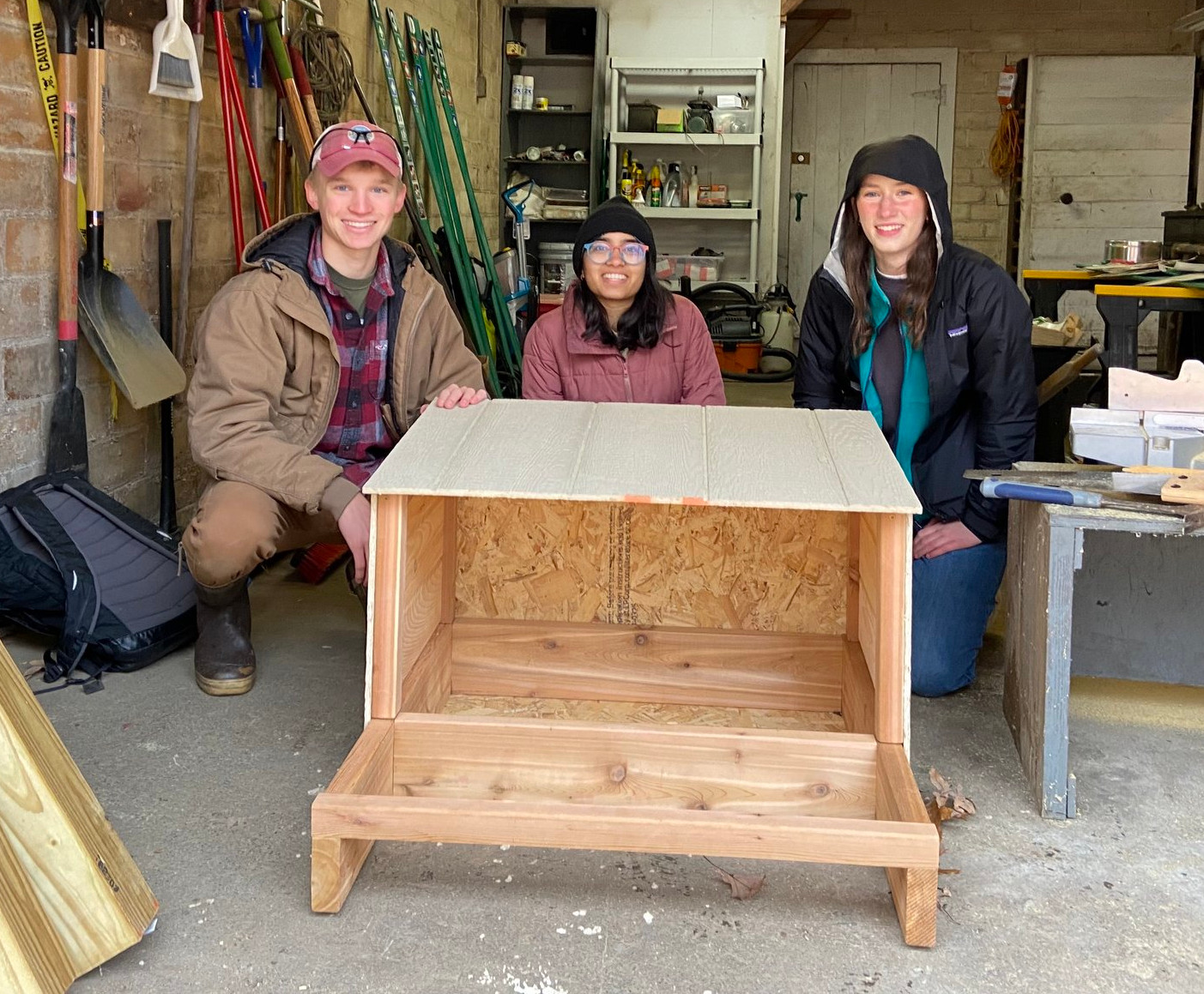 Three people kneel around a wood nest box in a workshop.