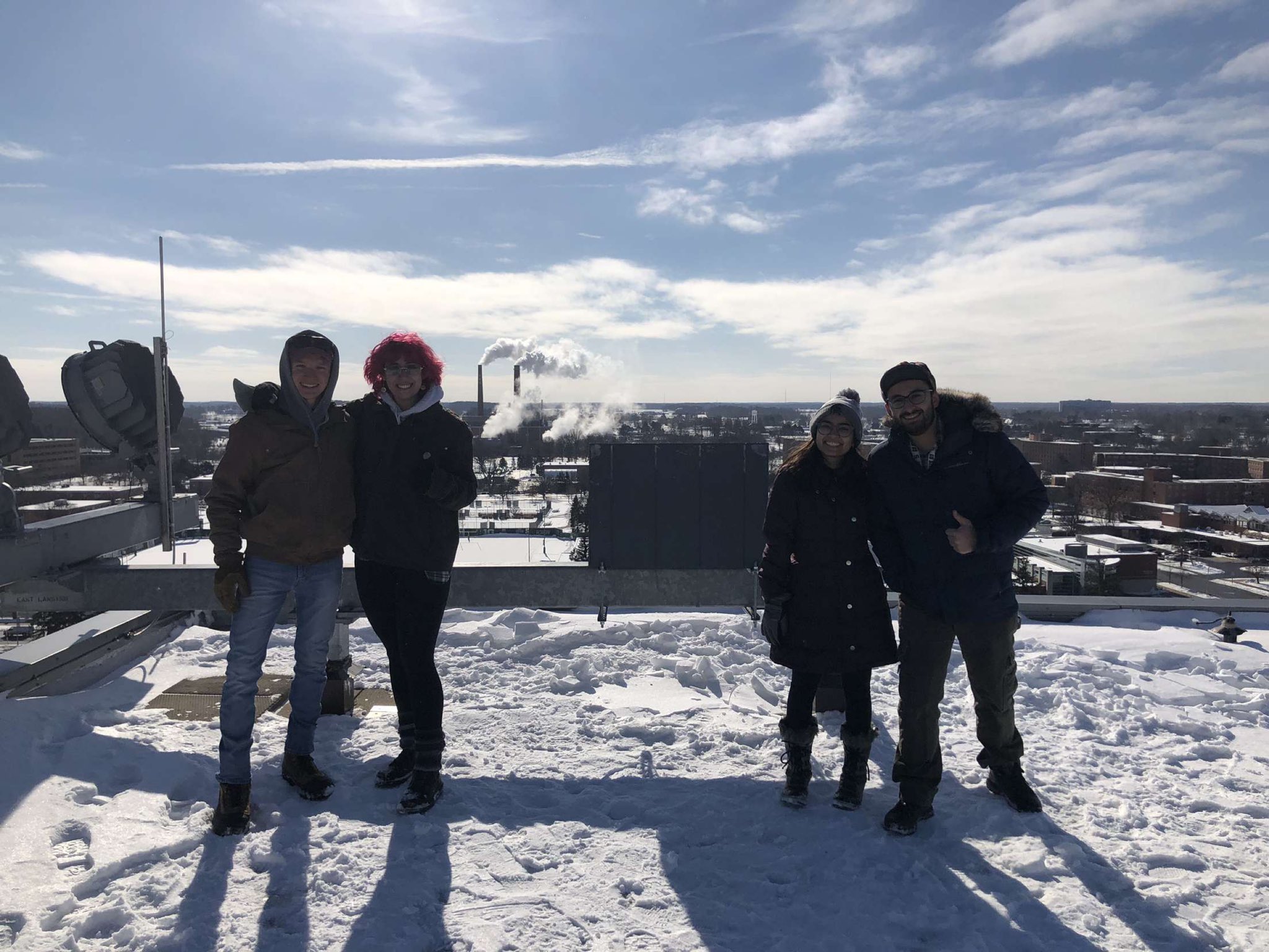 Four people standing on a snow-covered roof with blue sky and clouds.