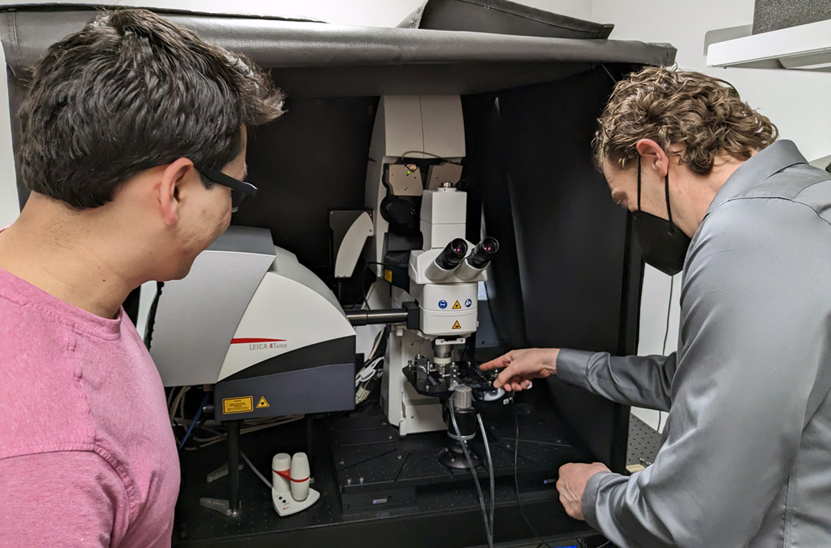 : A photo shows Bryan Smith (right) and Axel Schmitter-Sánchez (left) talk about a microscope in the background of the image. The microscope looks similar to one you might find in a high school science class: it has the familiar two eyepieces above a stage where a sample sits. But this microscope is powerful enough to reveal biological processes within living cells.