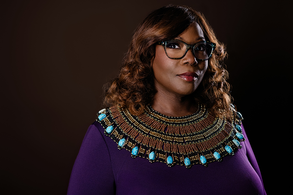 A studio portrait of a woman wearing purple with a beaded collar necklace and a black background.