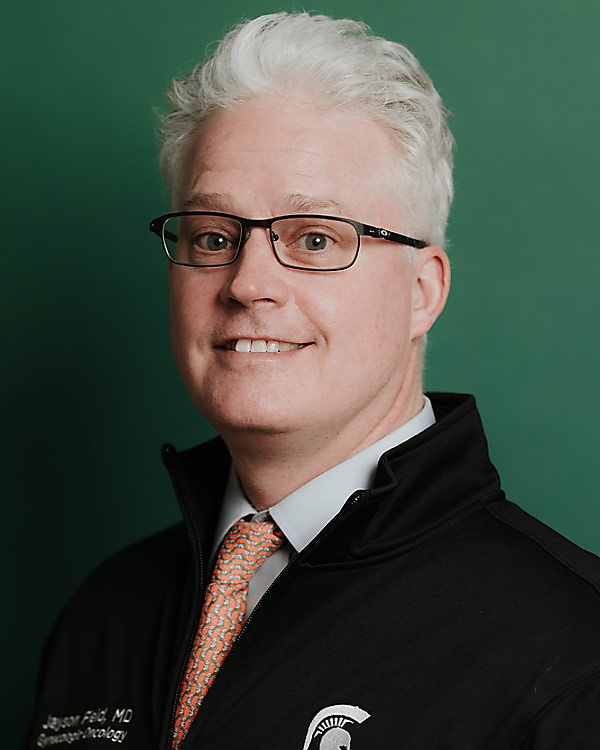 Jayson Field wearing a Spartan fleece over a shirt and tie. He is smiling with grey hair and black-framed glasses