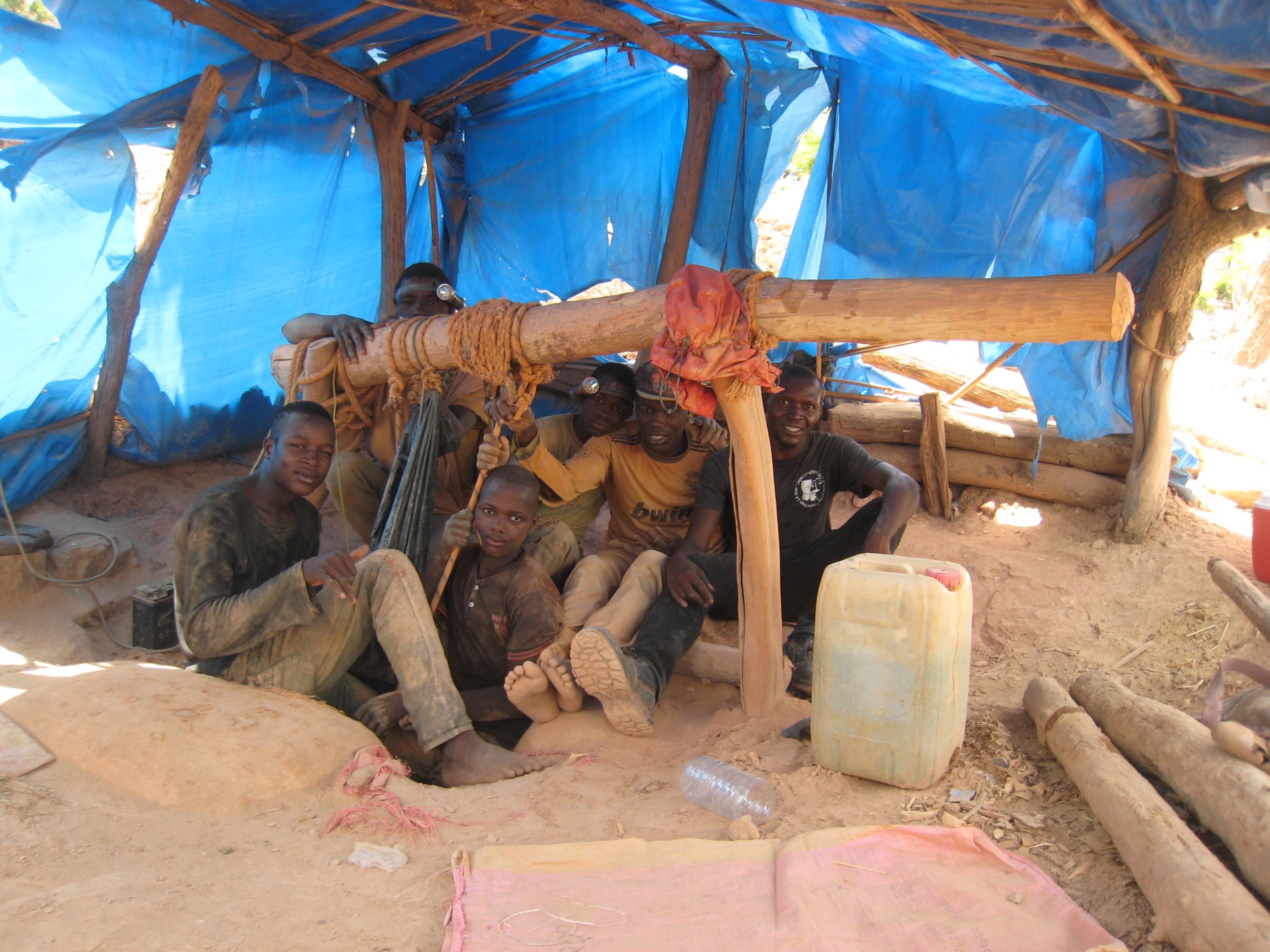 Miners pose for a photo near a dug-out mining pit under a blue tarp held up by a frame made from logs and branches.