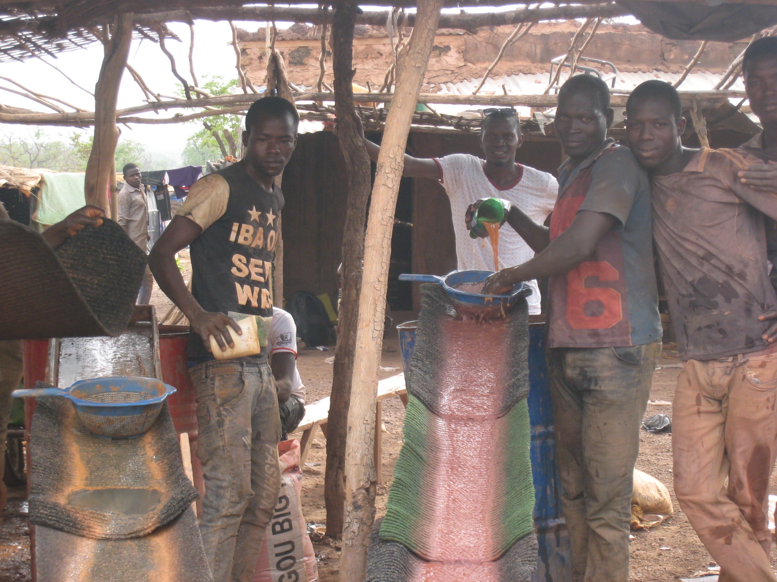 Miners pose for a photo while one pours an earth-toned liquid through a strainer and down a sluice to separate out the fine-scale gold-laden sediment from larger soil particles.