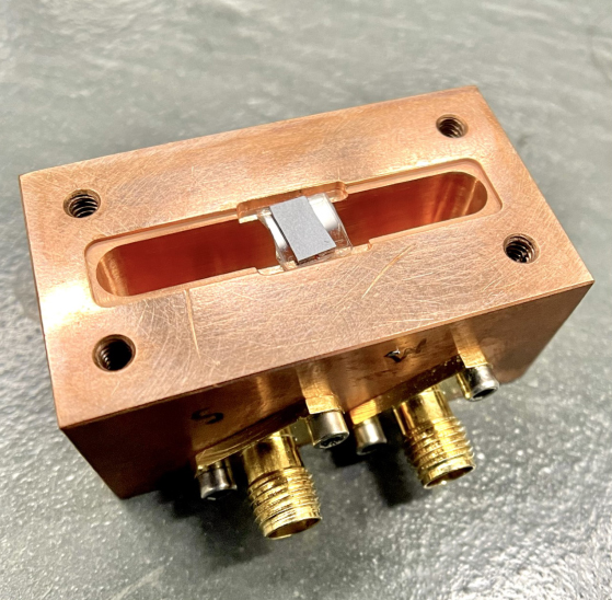 A photograph shows an experimental apparatus used by the Laboratory for Hybrid Quantum Systems at Michigan State University. A small, gray chip containing the quantum bit and surface acoustic wave resonator rests in the middle of the image on top of a small clear insulator. The stack sits on a larger copper-colored holder with gold-colored input/output ports.