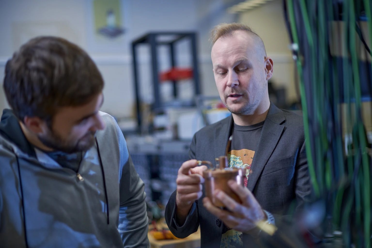Johannes Pollanen stands in the center of the frame holding a copper-colored cylindrical apparatus for holding qubits with Joe Kitzman looking on from the left. The two stand in a laboratory space with shelving and equipment in the background. Blue and green cables run down the right side of the photograph in the foreground.