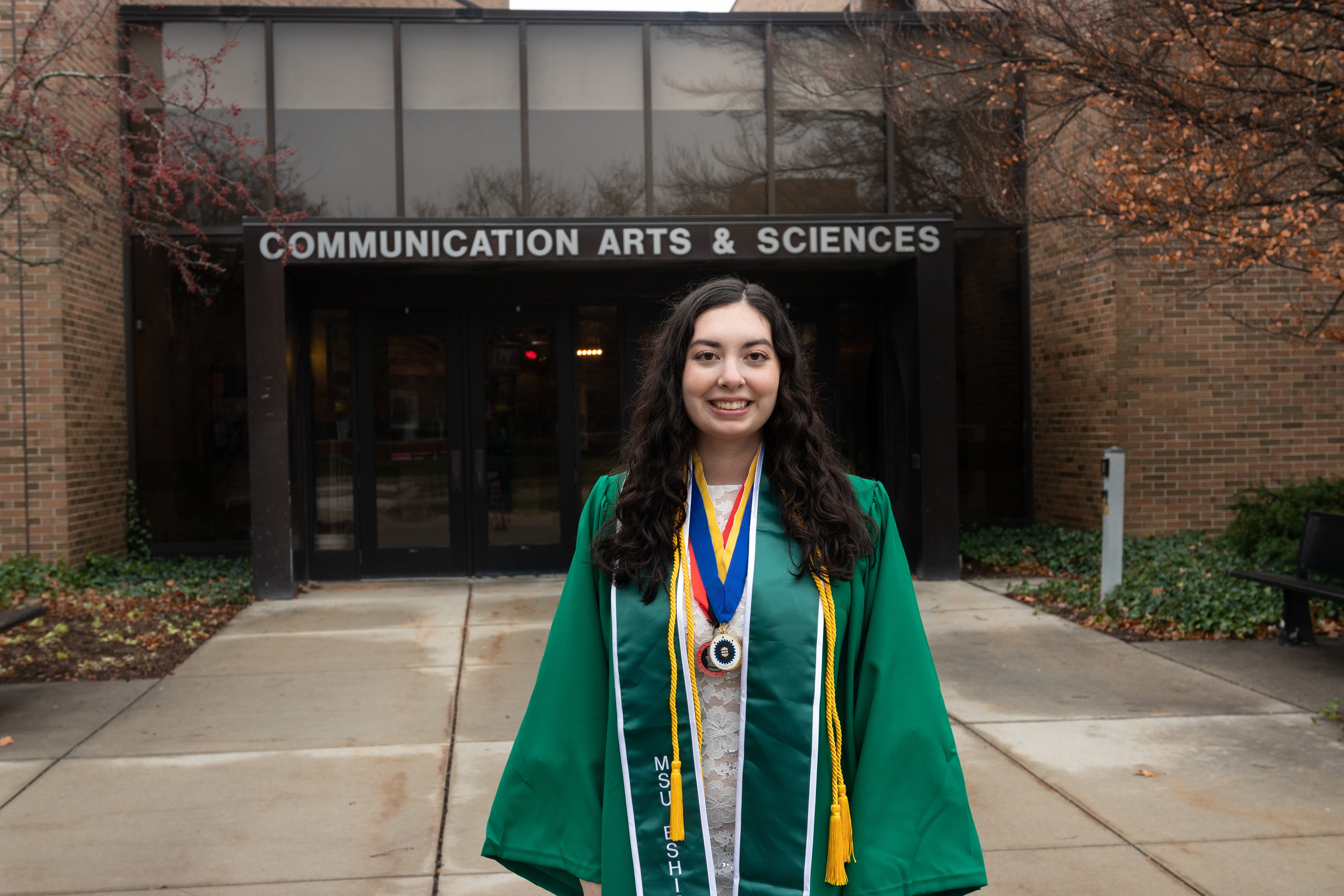 Gabrielle Sanchez in front of the Communication Arts and Sciences' building in green graduation regalia.