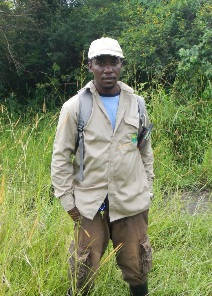 Sam Ayebare wears a hat and backpack and stands in a field with long grass and trees behind him. 