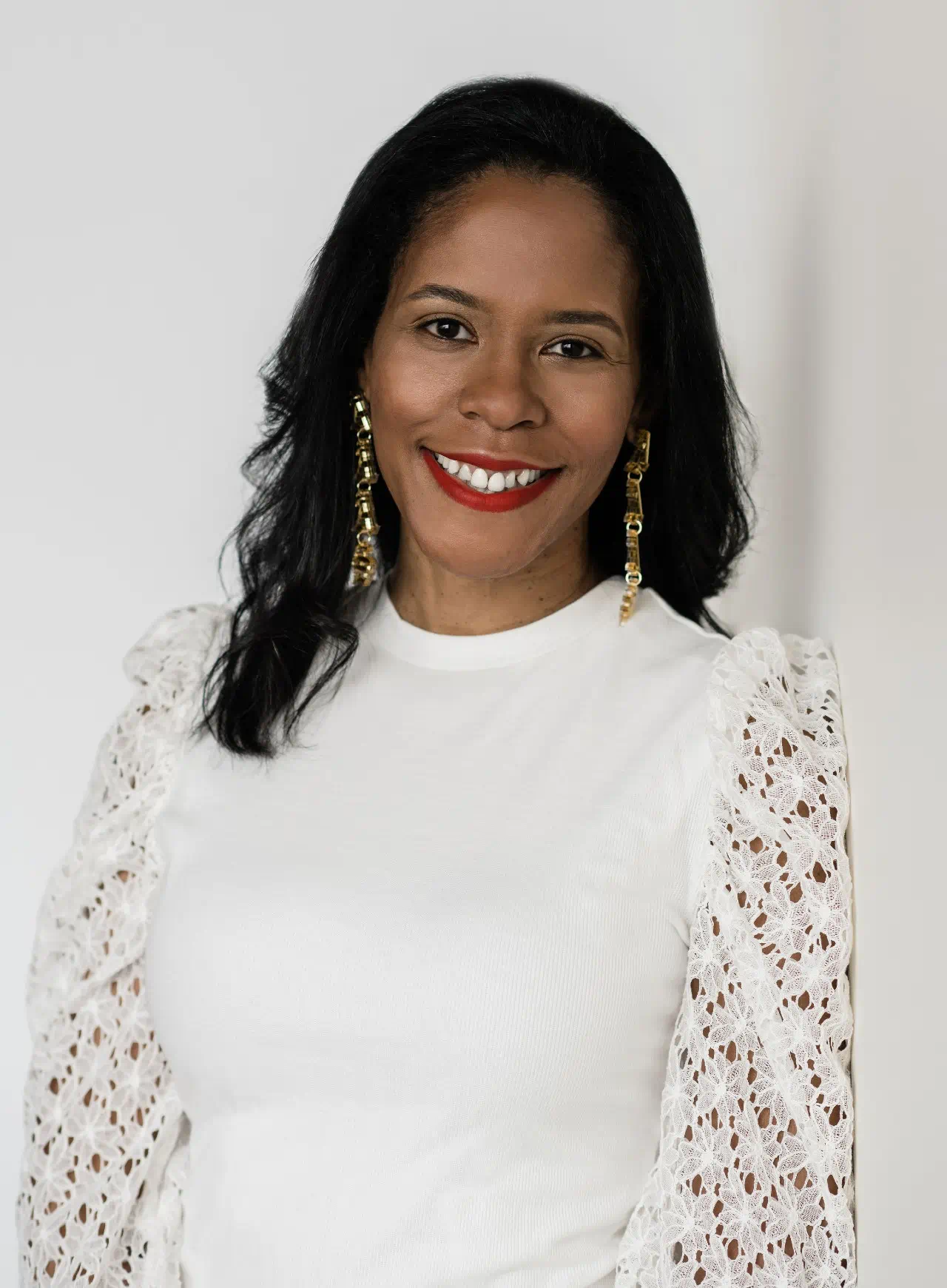  Headshot of Ruth Nicole Brown. She is smiling. She has on red lipstick, gold earrings, and is wearing a white shirt while being photographed in front of a white background. 