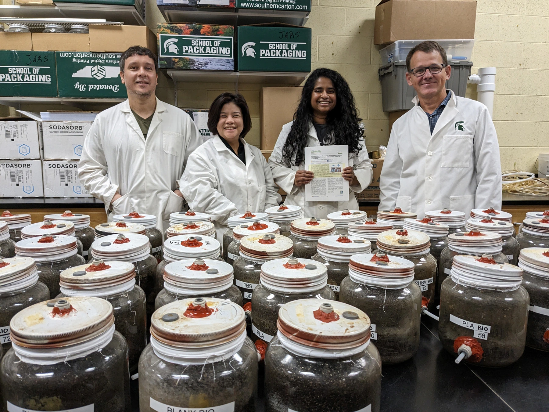 Anibal Bher, Wanwarang Limsukon, Pooja Mayekar and Rafael Auras stand in their lab wearing white lab coats. There’s a table in front of them holding dozens of bioreactors — clear glass jars sealed tightly with lids and filled with compost.