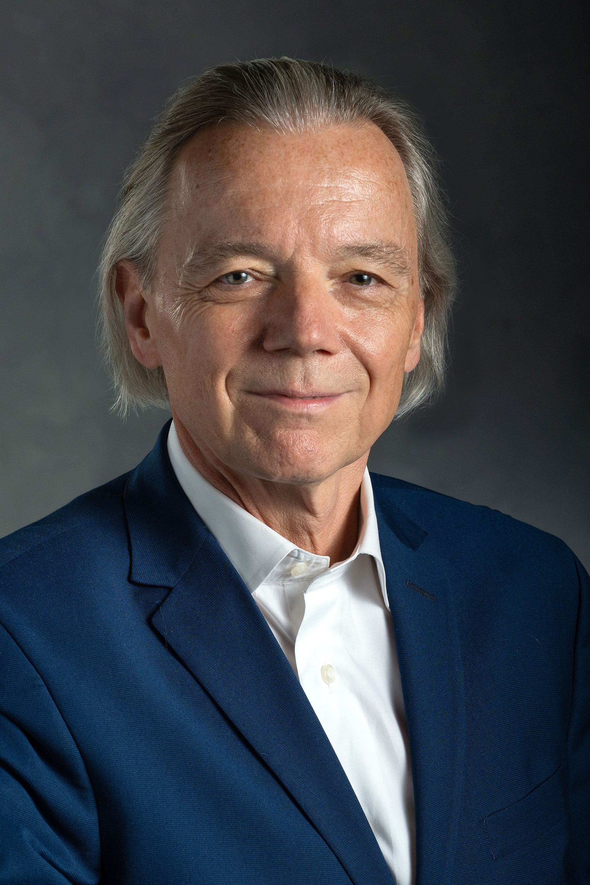 A headshot of Johannes Bauer posing in front of a gray background. He is wearing a navy suit jacket and white dress shirt