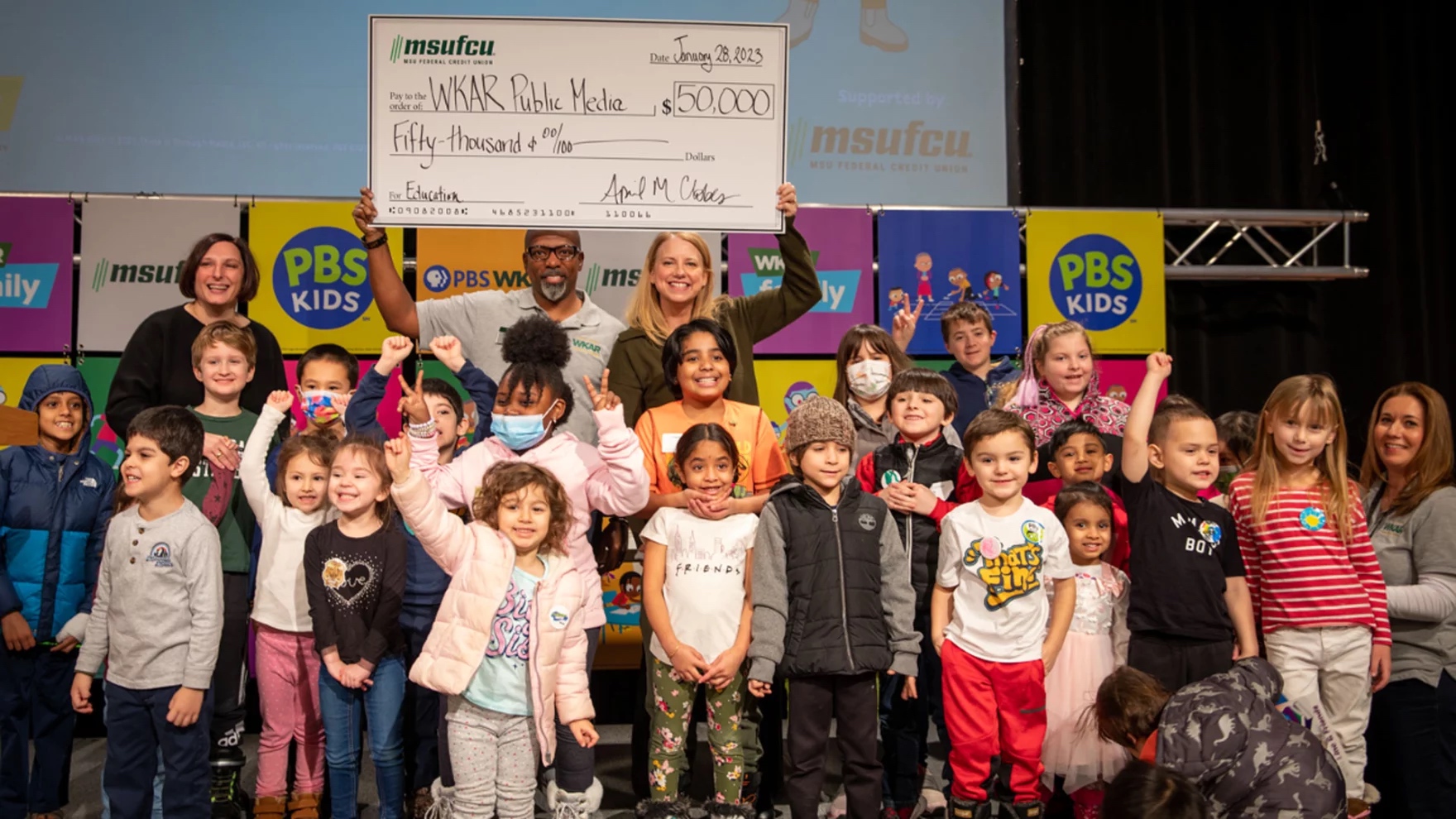 MSUFCU presents a check for $50,000 at the PBS KIDS Day with WKAR celebration on Jan. 28, 2023. April Clobes and Susi Elkins of MSUFCU (back row left and right) and Shawn Turner and Julie Sochay of WKAR (back row center and front row right) pose with just a few of the hundreds of children attending the event.