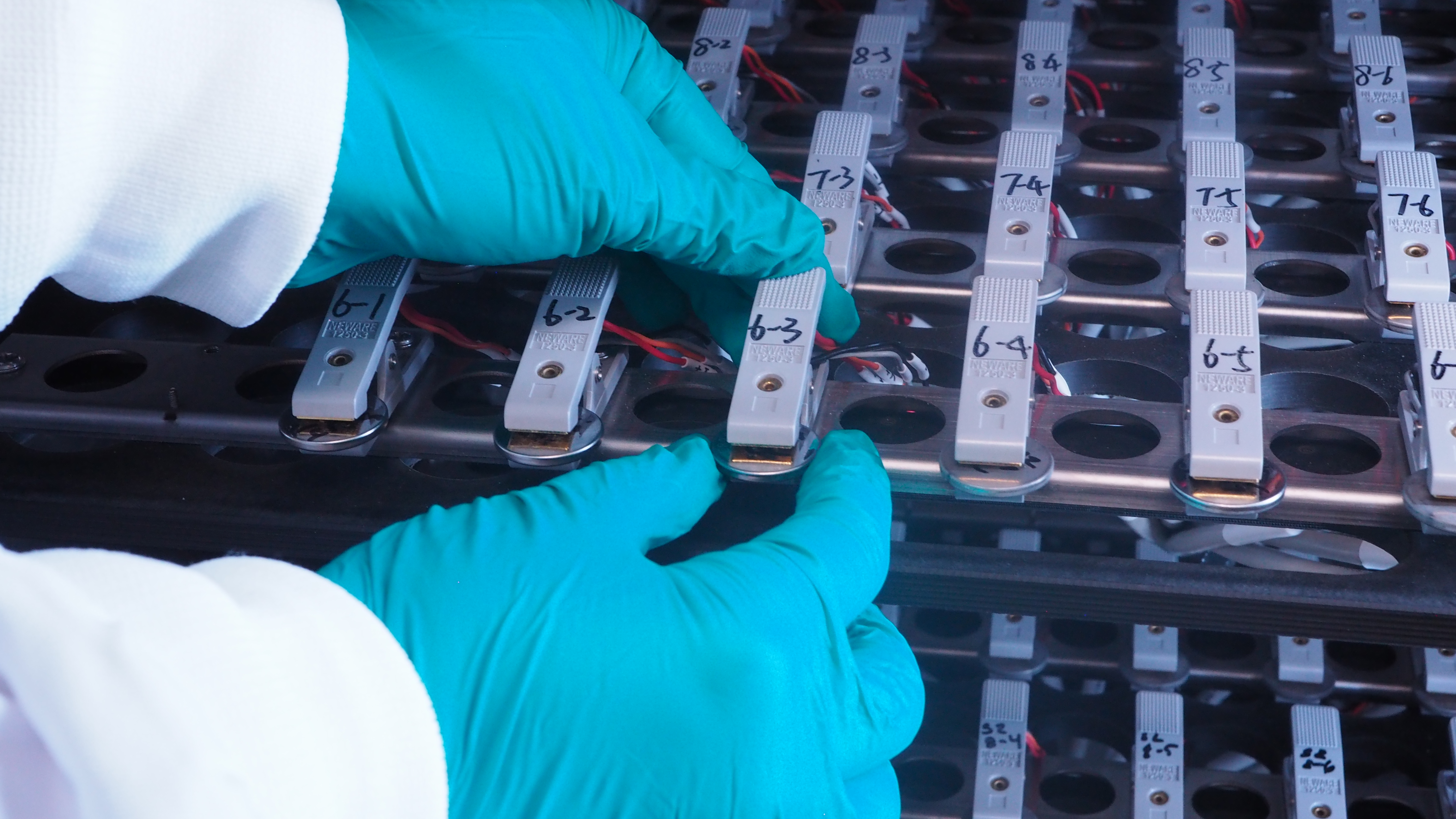 A look inside a battery cycler where rows of dozens of coin-shaped batteries are held in place by clamps bearing sample numbers.