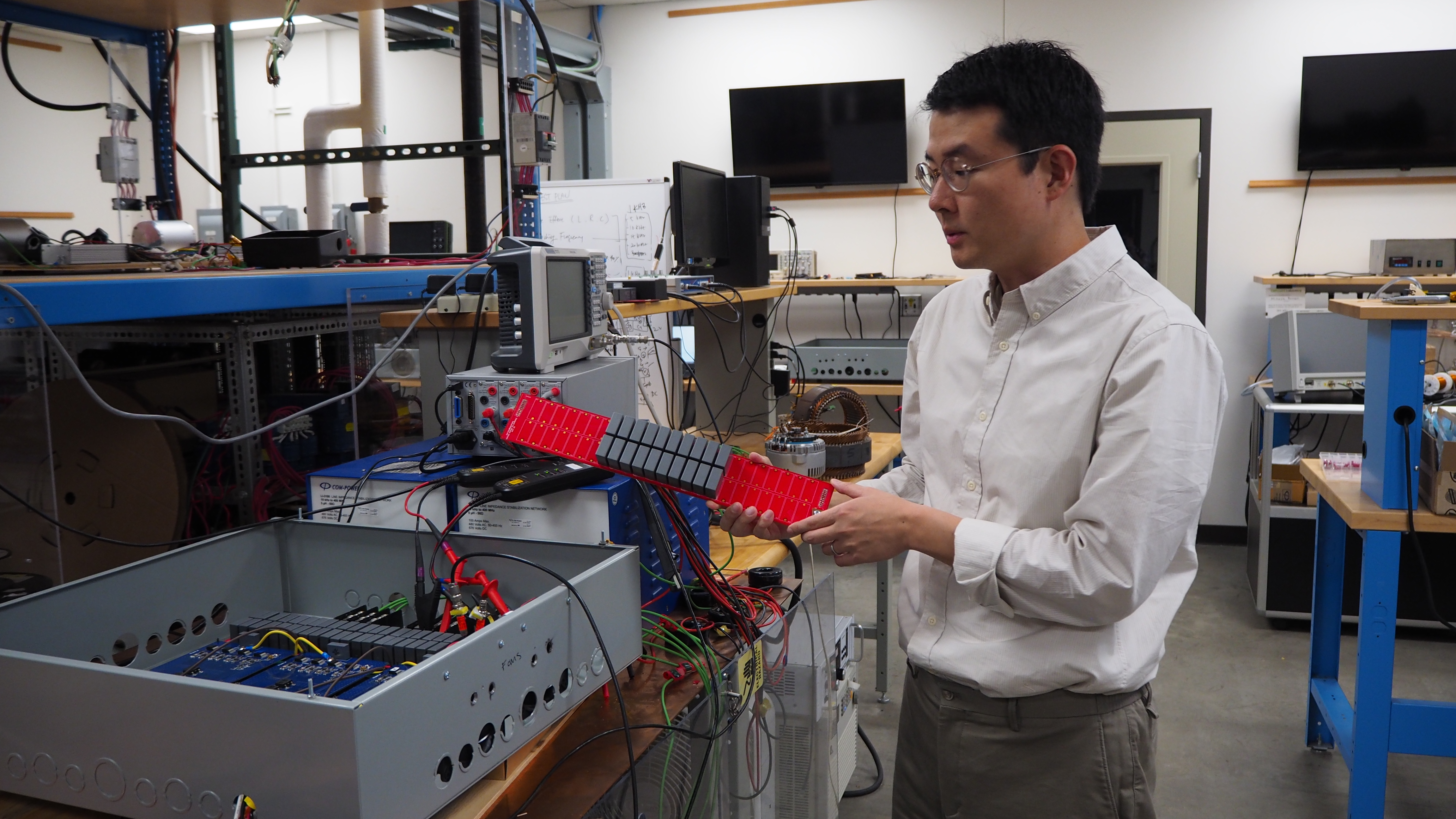 Woongkul “Matt” Lee stands next to an open metal box, about the size of a large sheet cake, filled with wires and electronic components. He holds one of those components — a narrow red board with about 20 small gray capacitors mounted to it — for a better look.