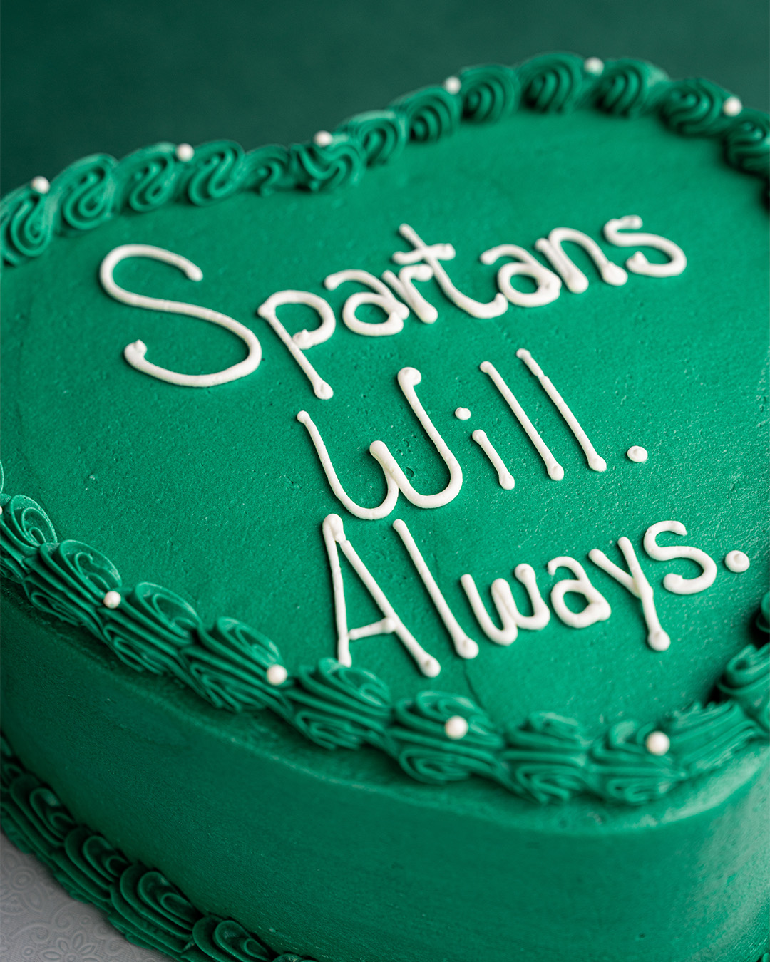 Heart-shaped cake with green frosting, white writing in icing on top that reads Spartans Will. Always.