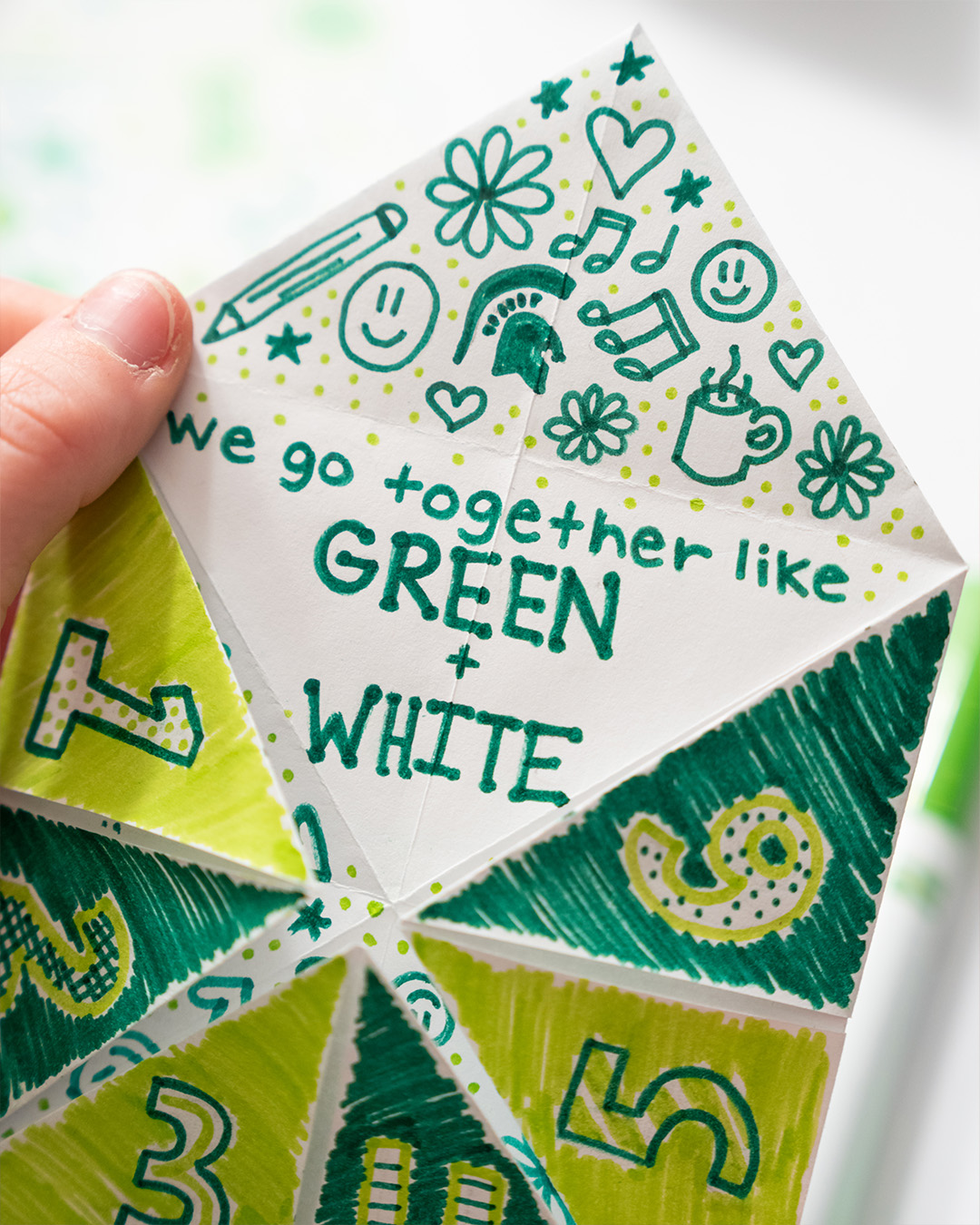 Origami fortune teller decorated with green marker designs; one flap open reading we go together like green + white