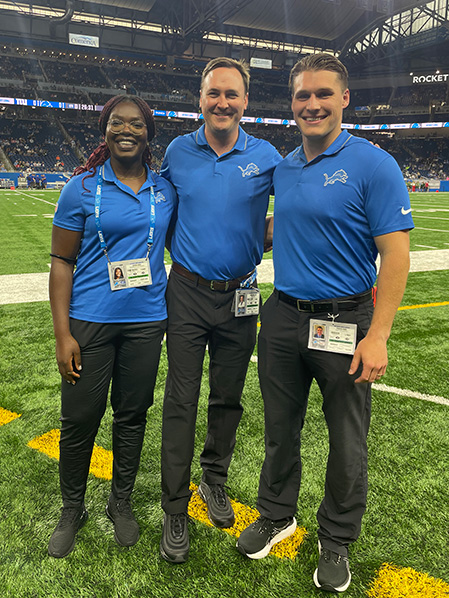 Tamarandobra “Dobra” Ogeh poses with two men on the sidelines at Ford Field, wearing a blue Detroit Lions polo shirt