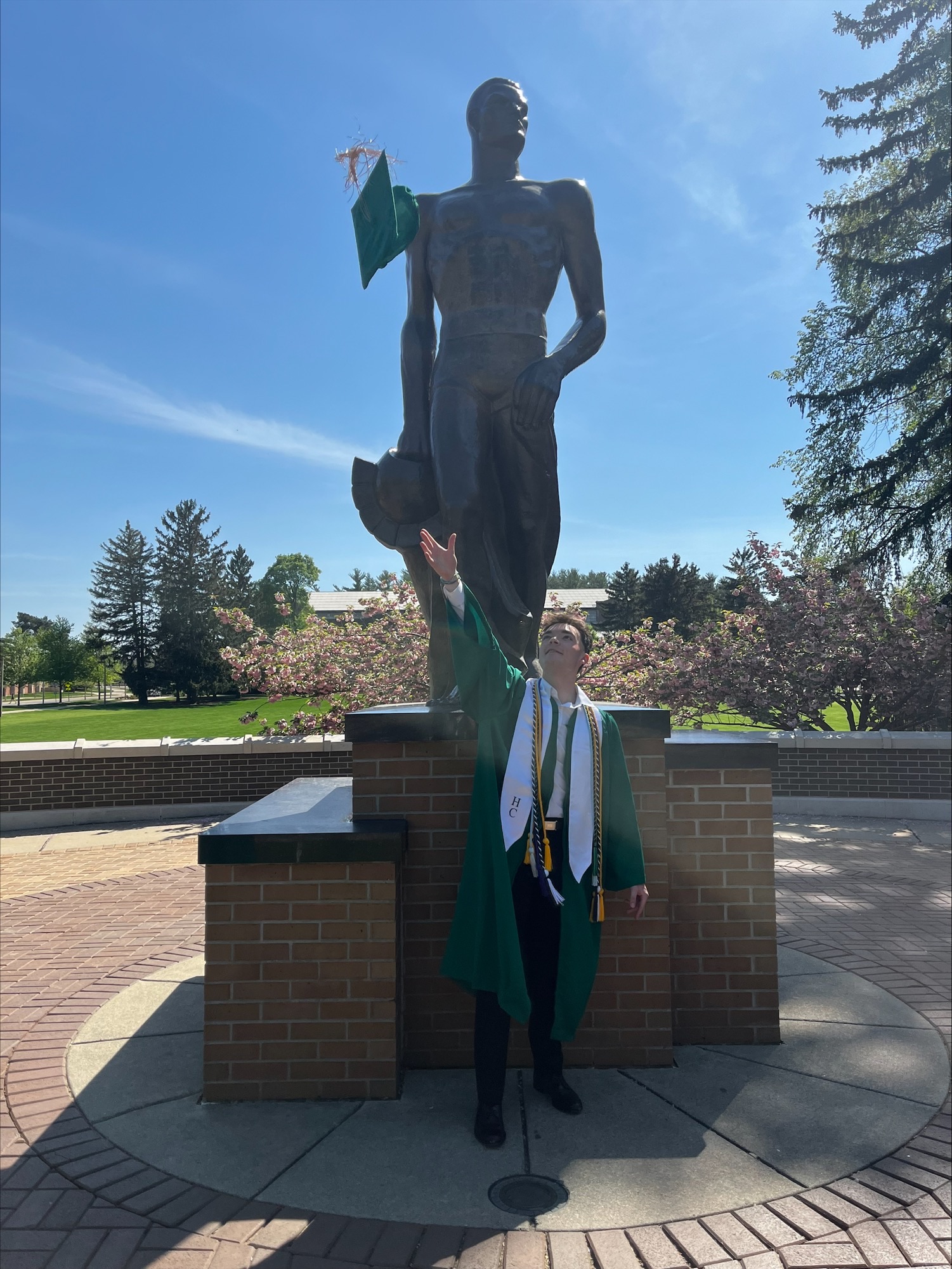 Carlos Conrad in his green graduation robe standing in front of The Spartan statue.