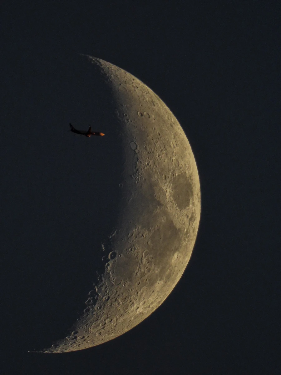 A photograph taken with the help of the MSU Observatory’s telescope shows an airplane flying in front of a waxing crescent moon. The plane is in front the dark side of the crescent. Craters and smooth areas are detailed on the crescent’s bright side.