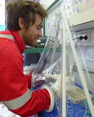 Dalton Hardisty works with scientific samples inside a glove bag, which is like a small, clear plastic tent designed to keep air from reaching the samples inside. Hardisty is able to handle the samples by placing his hands in large white gloves built into the side of the bag.