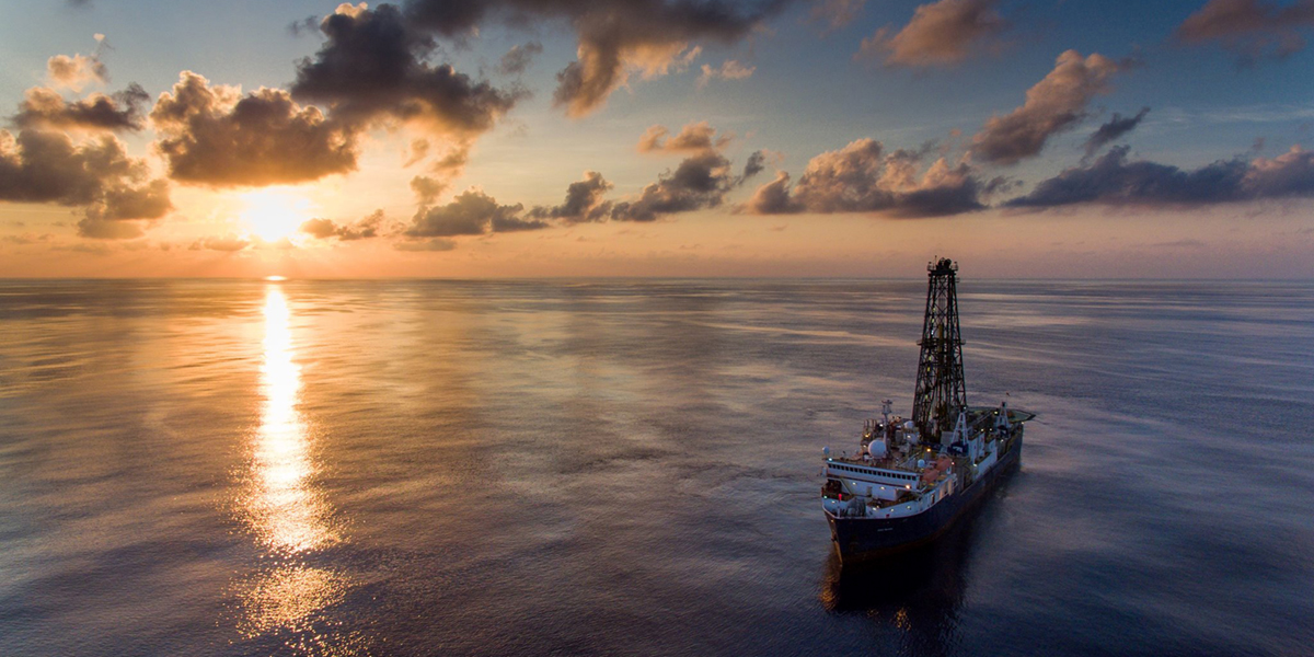 The sunset casts a streak of bright yellow reflections across the surface of an otherwise blue South China Sea. A large ship with a nearly 150-foot drilling tower at its center floats to the right of that streak, under a dusky sky with wispy clouds.