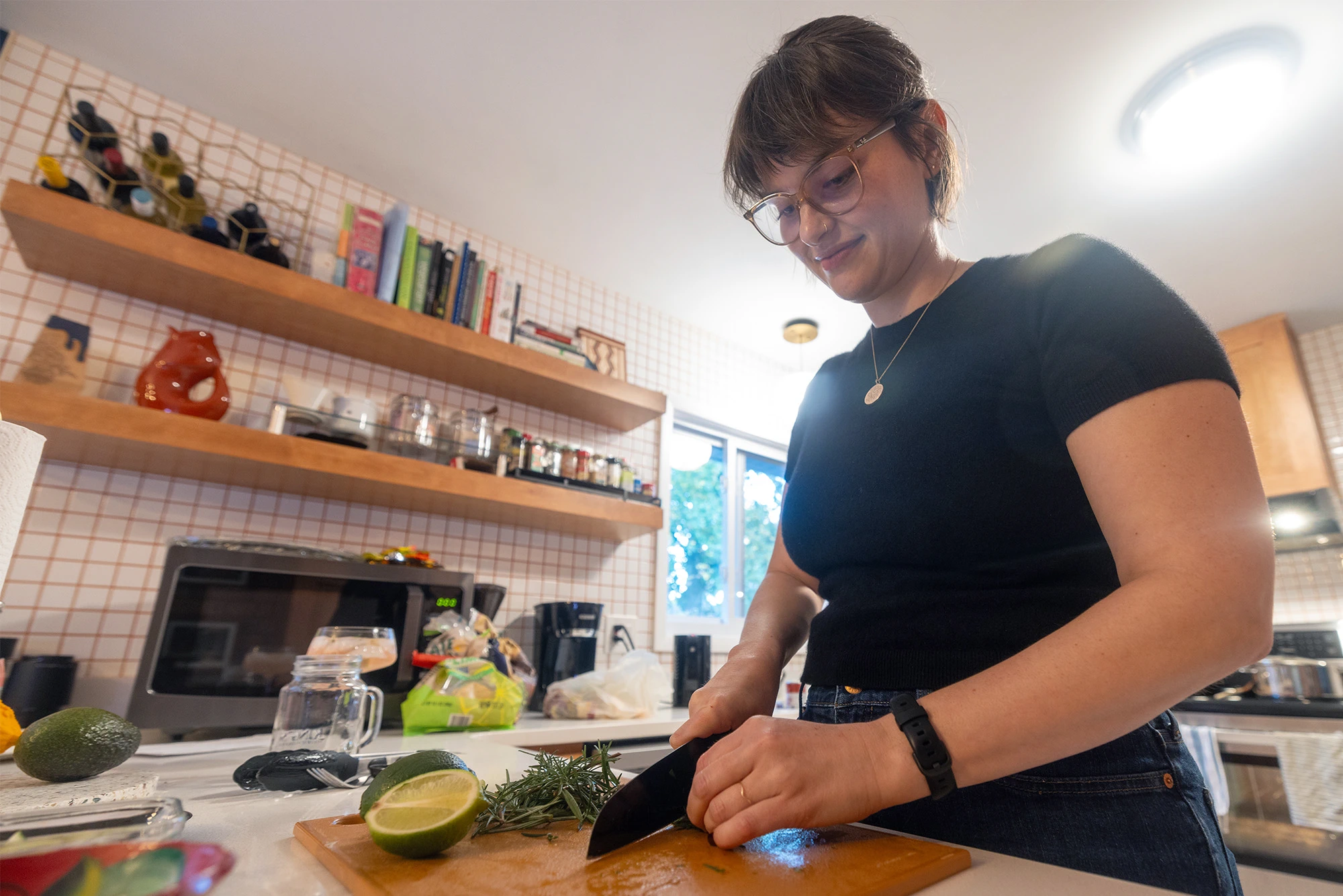 Megan Dean wearing a black shirt and glasses uses a knife to cut herbs on a cutting board in a kitchen setting.