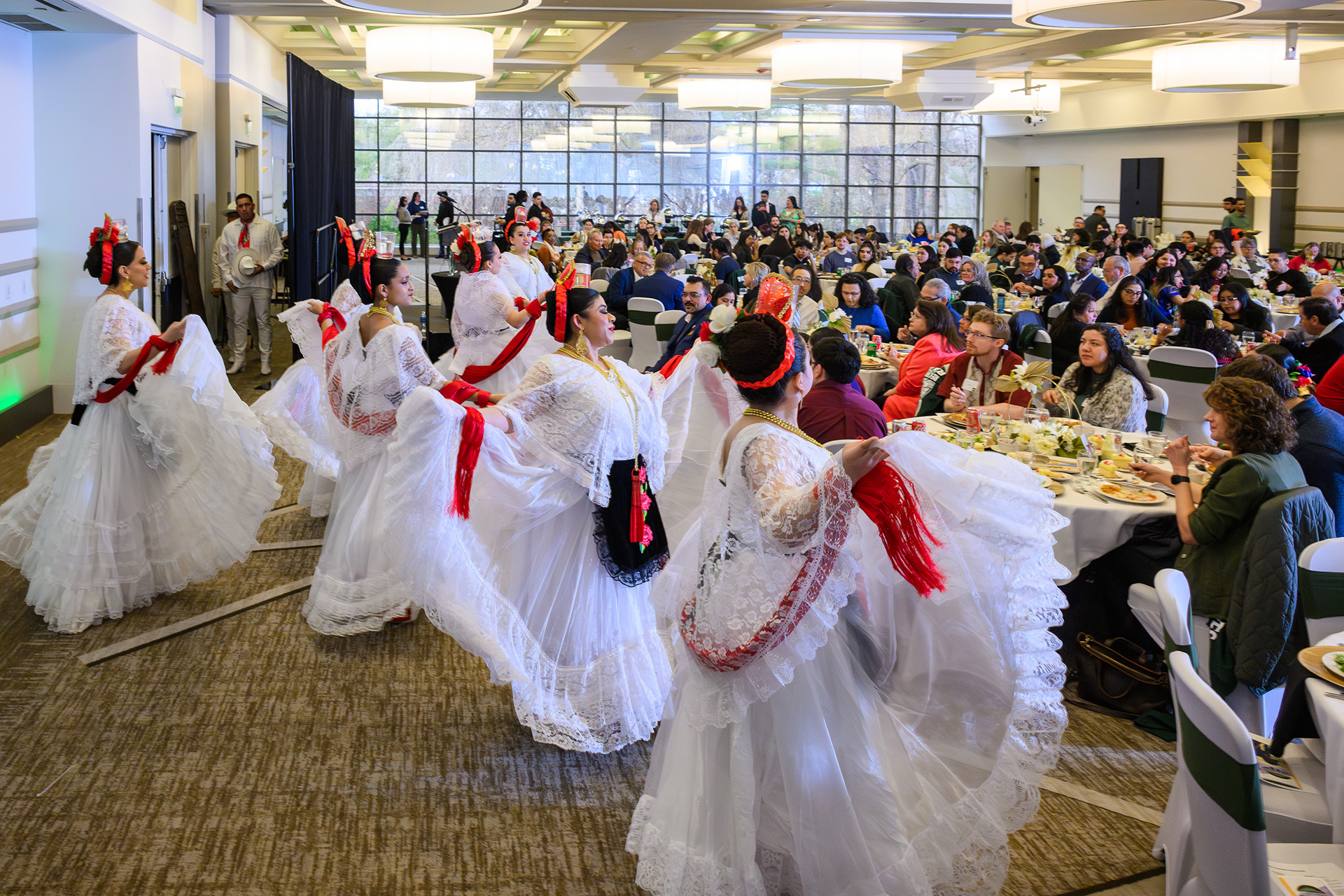 six dancers wearing flowing white dresses with red accents perform in front of the full room