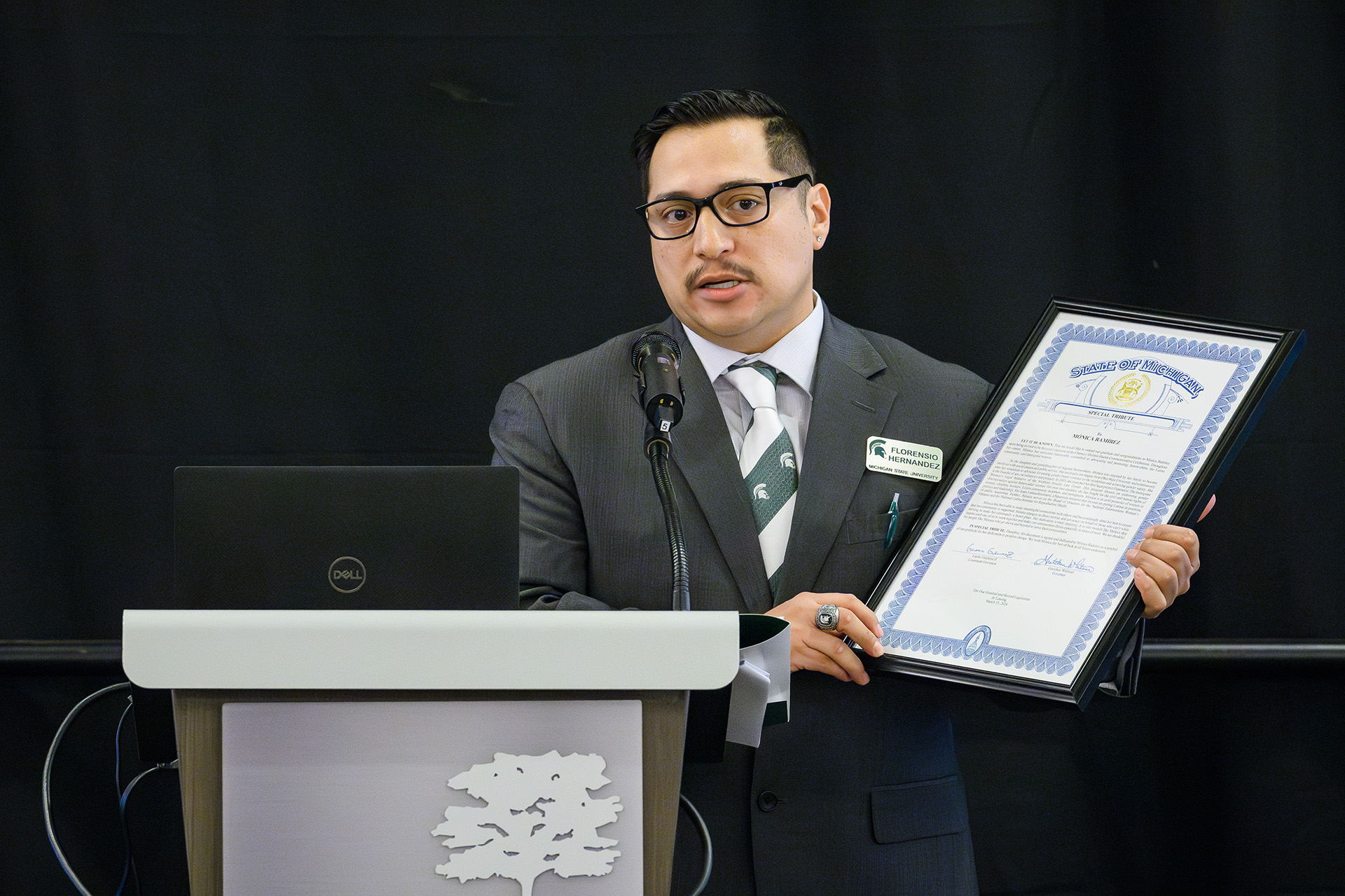 Florensio Hernandez speaks at the lectern holding a large framed Michigan state certificate recognition