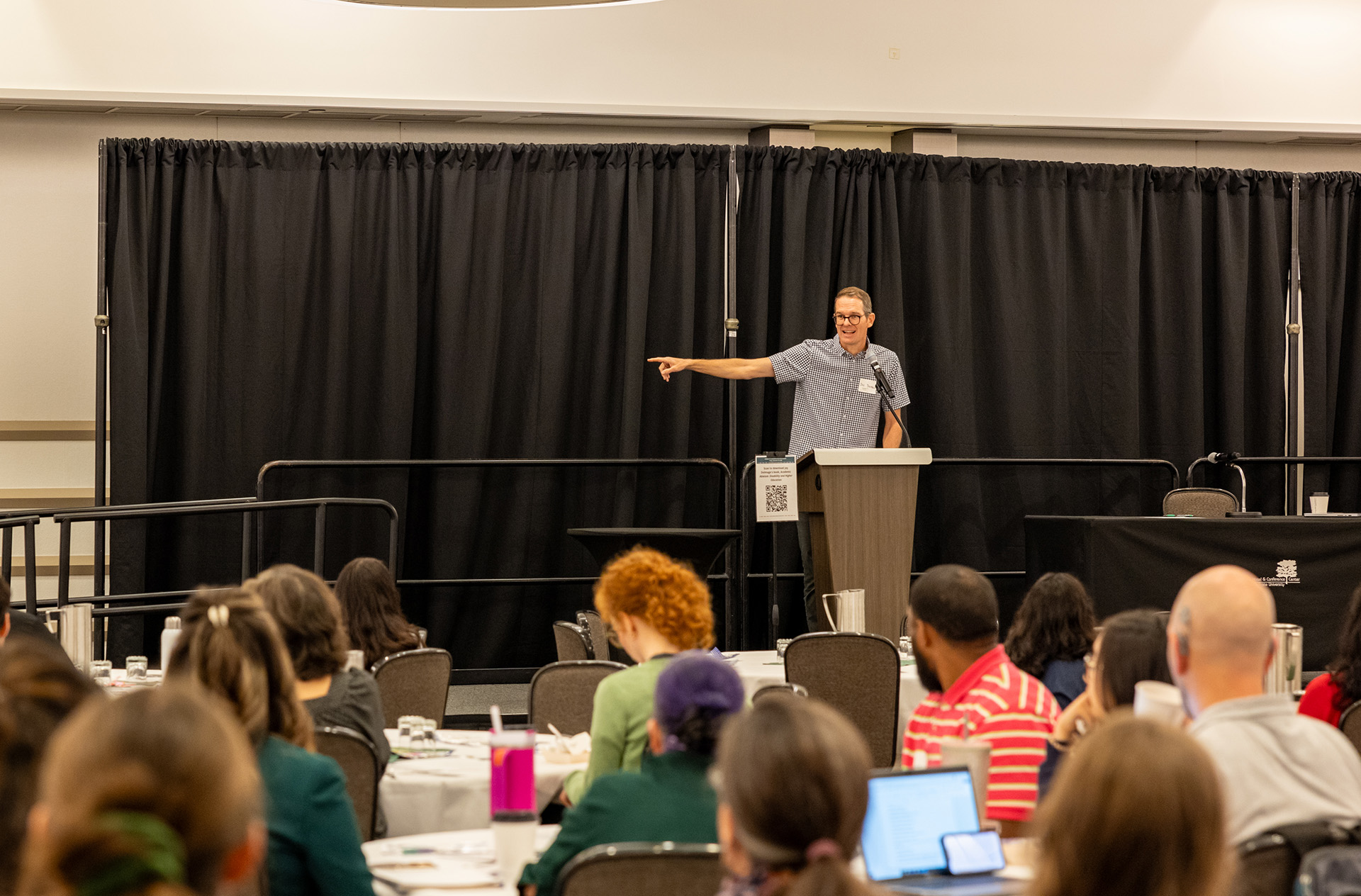 a speaker stands at the podium and points to the side in front of a room of participants