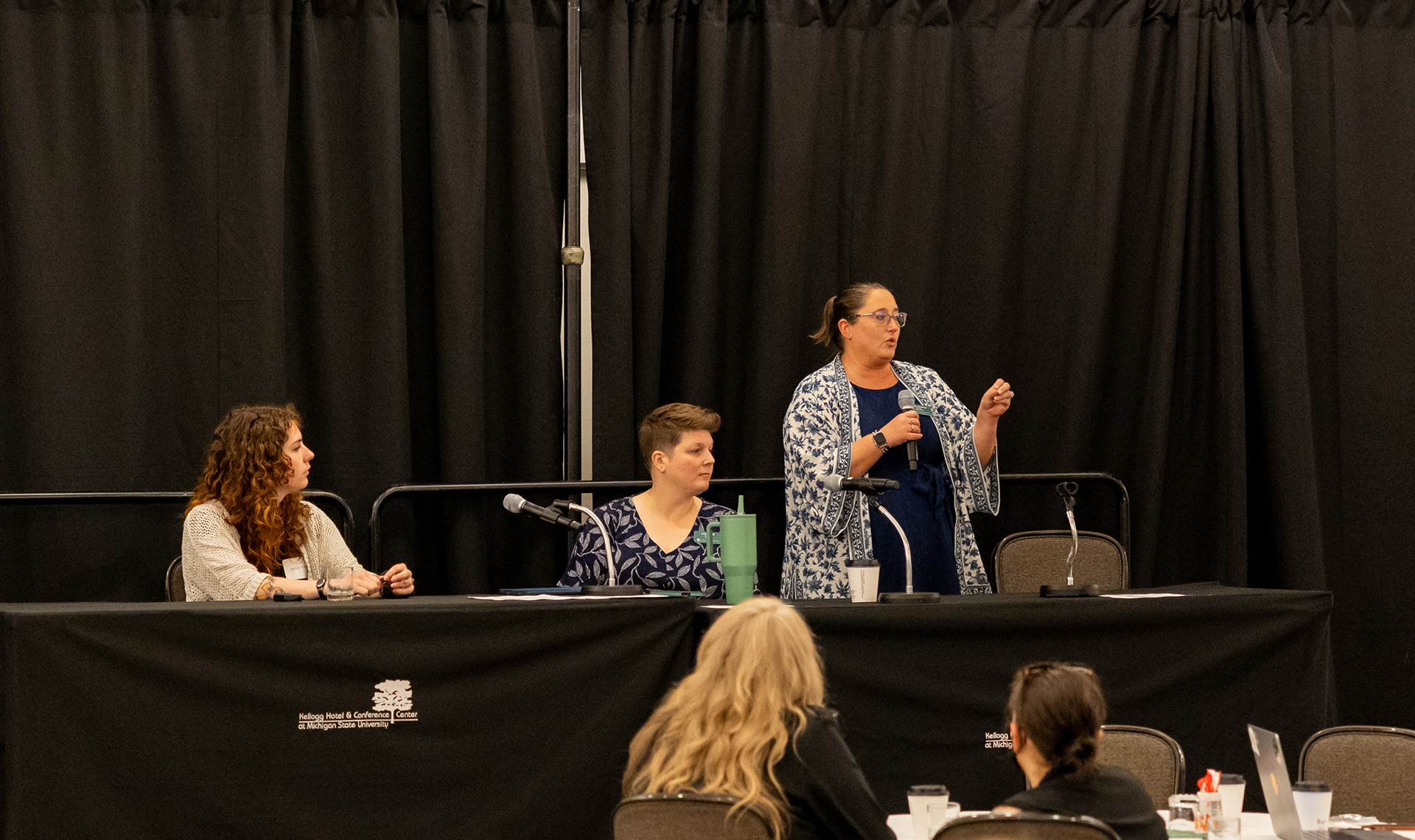 three panelists at a table, with one standing and speaking into a mic