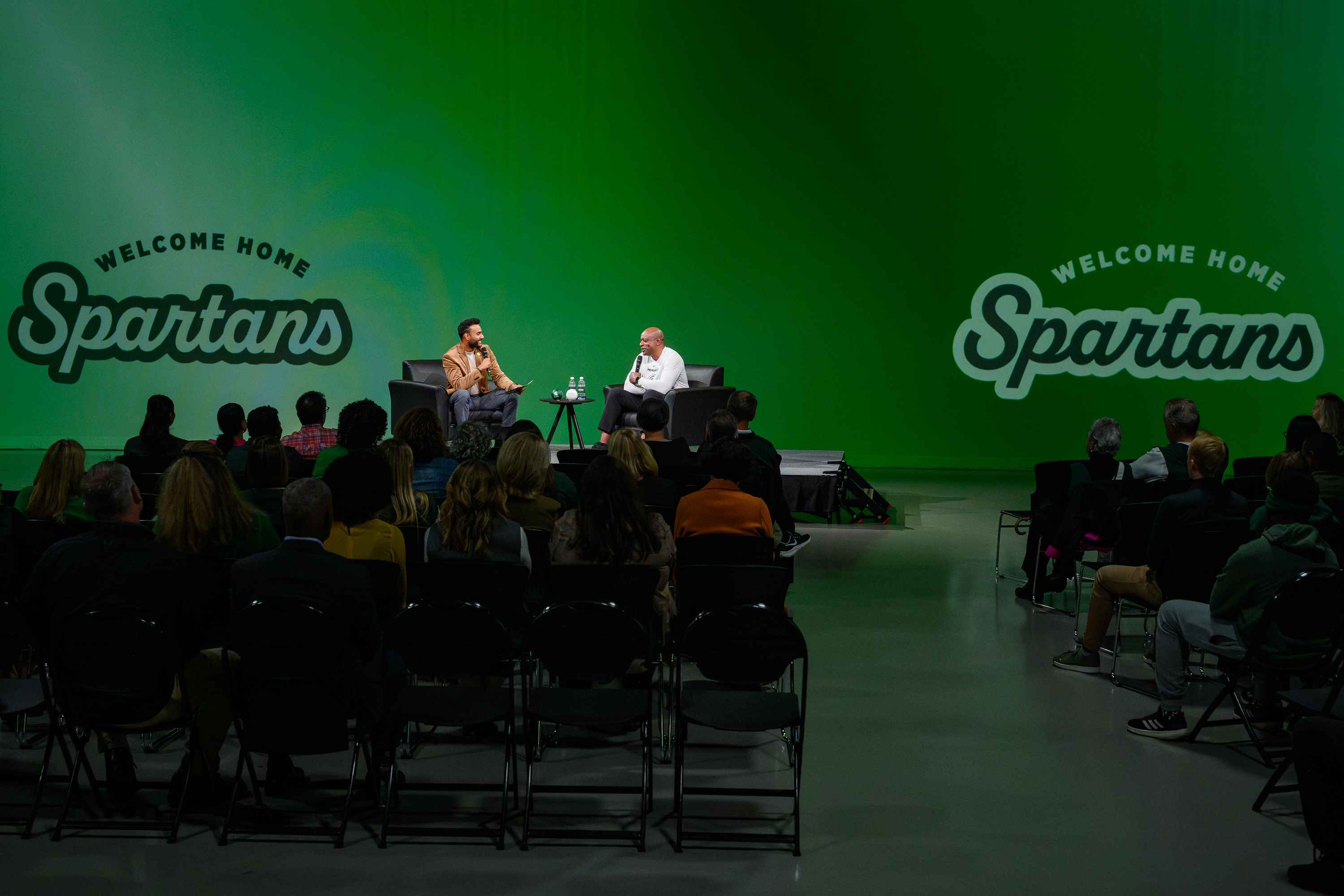 A crowd faces two people on the stage with a green background.